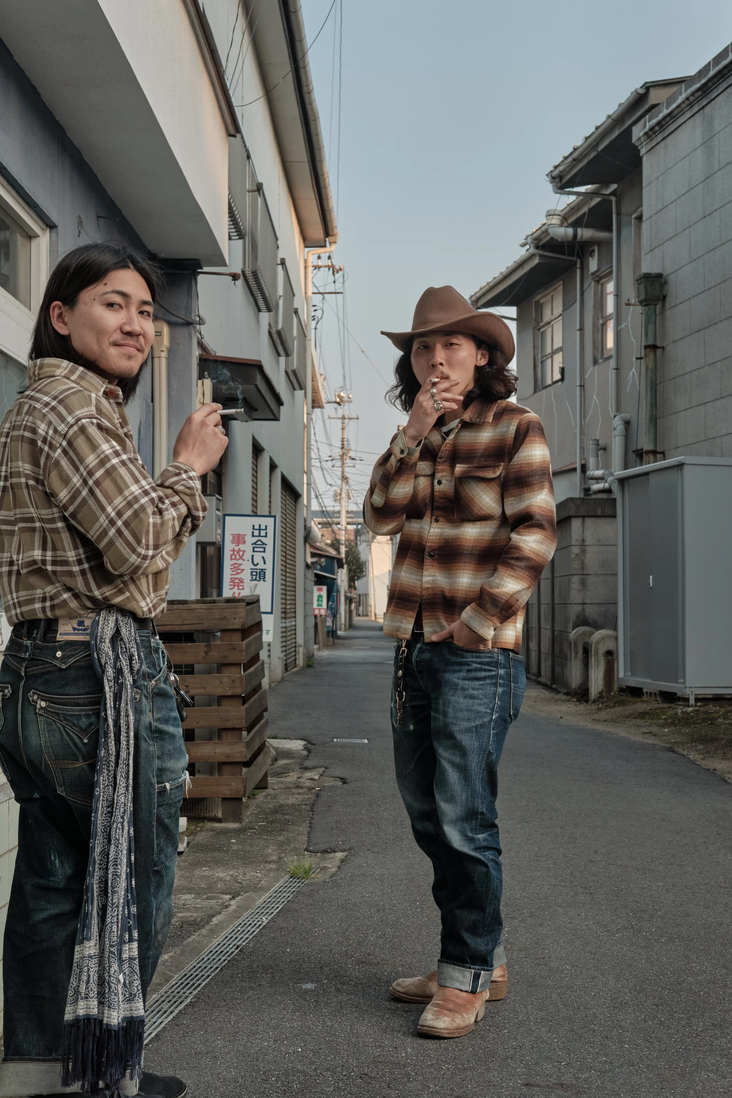 Two young men standing on a narrow street in a Japanese neighborhood, one smoking and the other looking at the camera, both dressed in vintage-style plaid shirts and jeans. Kojima, Japan.