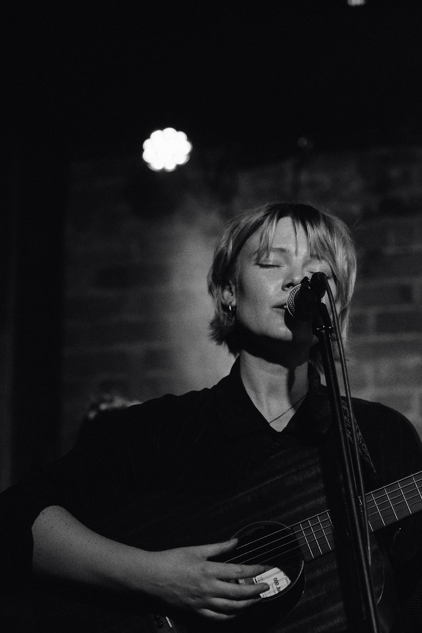 A woman with short hair and earrings playing an acoustic guitar and singing into a microphone on stage, eyes closed, in a dimly lit setting.