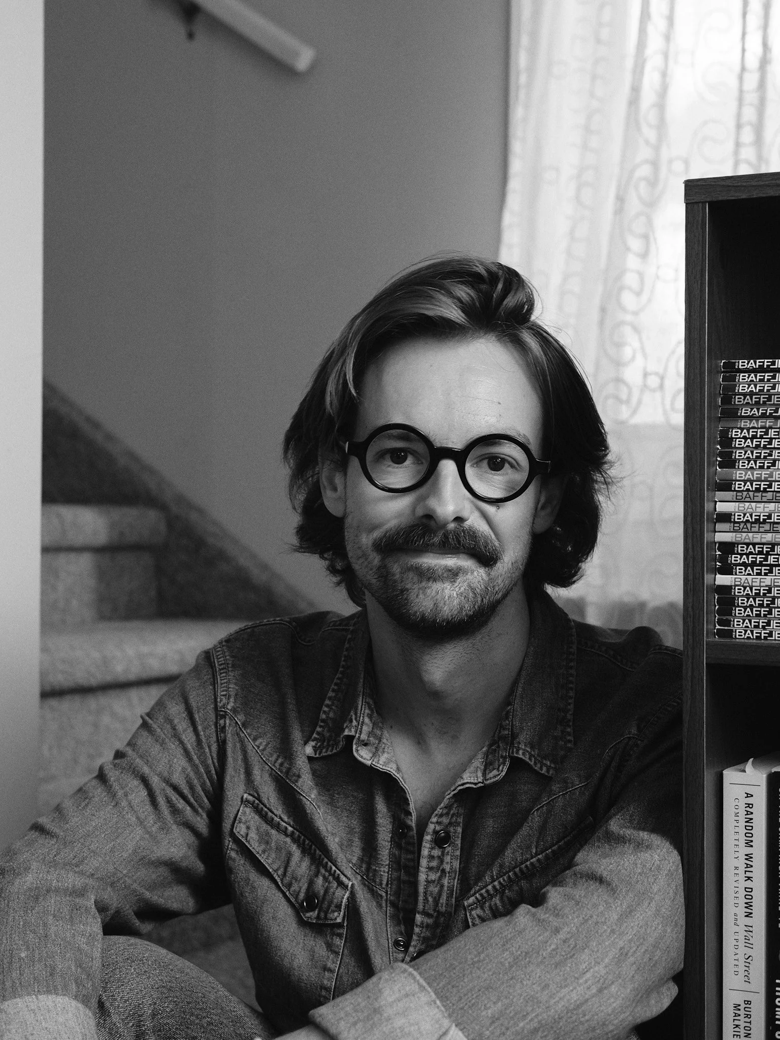 Black and white photo of a man with glasses, shoulder-length hair, and a beard, sitting indoors near a window with curtains, wearing a denim jacket. Jon Rode, photographer. Edmonton, Alberta.