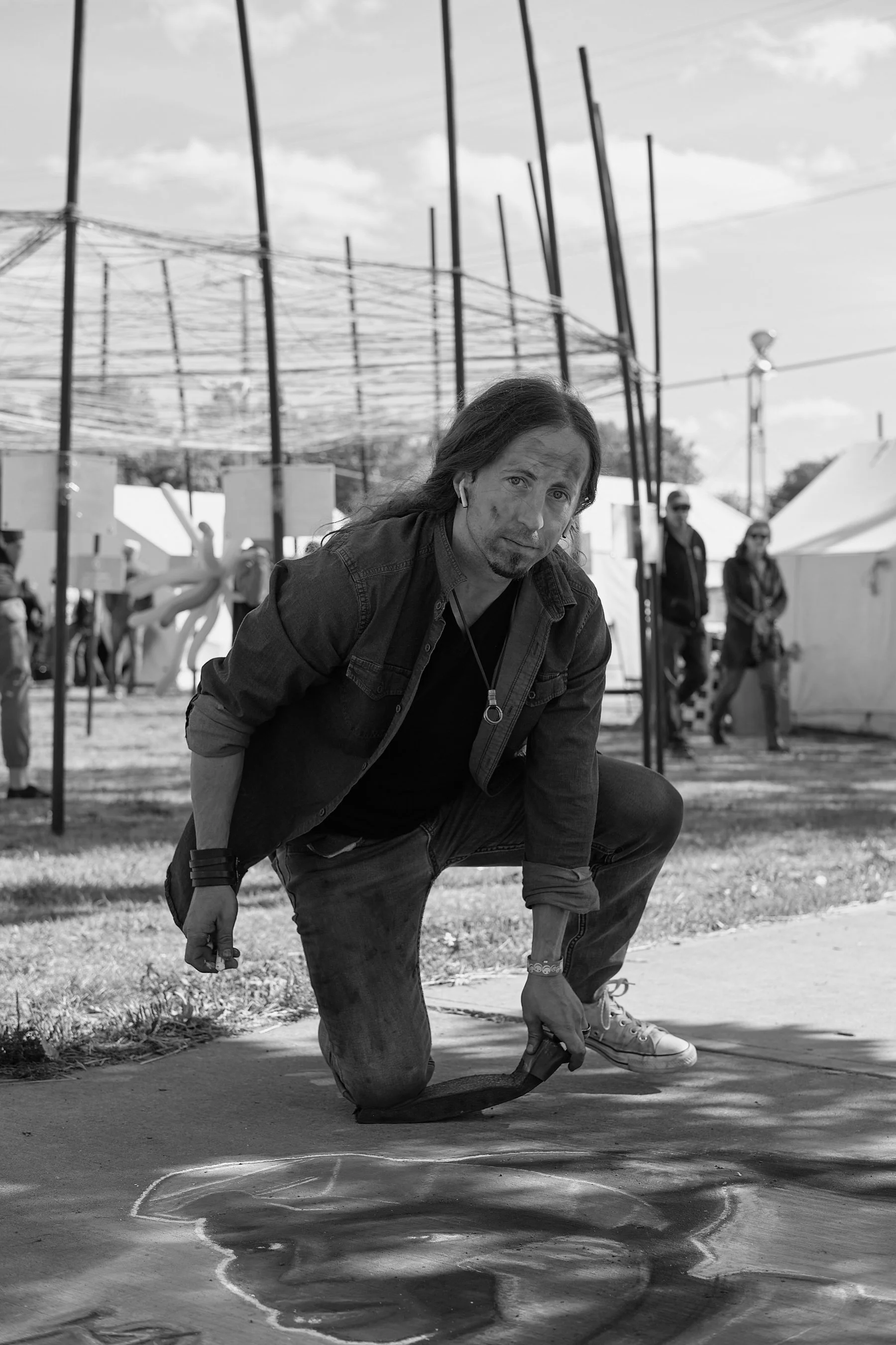 A man with long hair and a goatee, wearing a jacket and sneakers, is kneeling on the ground with one knee on the sidewalk and holding a skateboard. He has a serious expression and is looking at the camera. In the background, there are tents, people, 