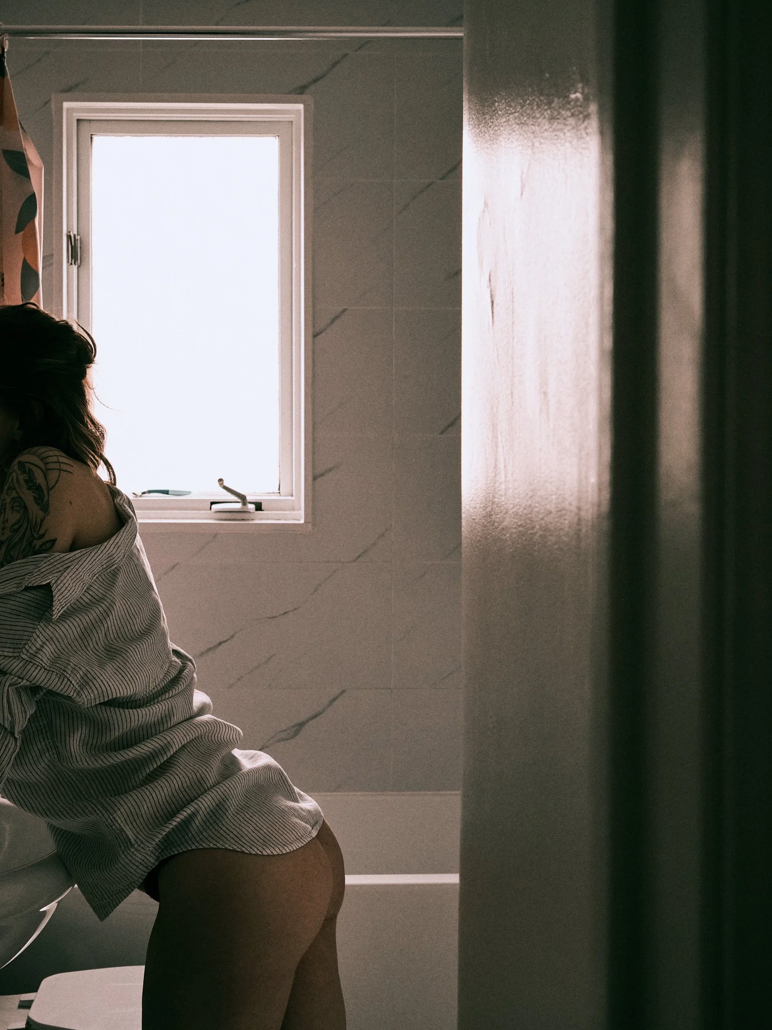 A woman in a striped shirt leaning into the mirror in a bathroom, viewed from the side, with a window in the background.