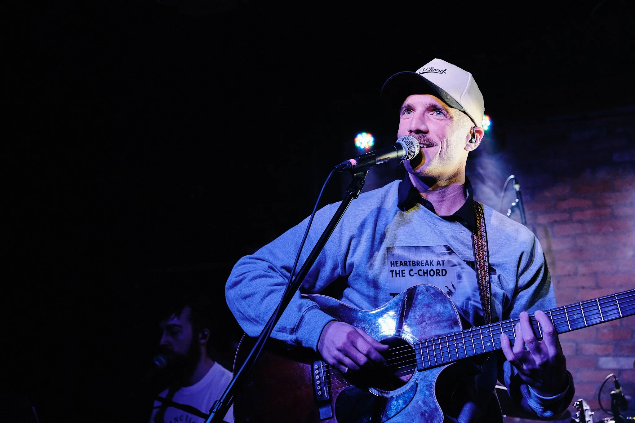 David Henneberg. Musician wearing a white hat and a grey sweatshirt that says "Heartbreak in the C Chord" playing guitar on a stage and smiling. Taken at Dive Bar in Edmonton, Alberta.