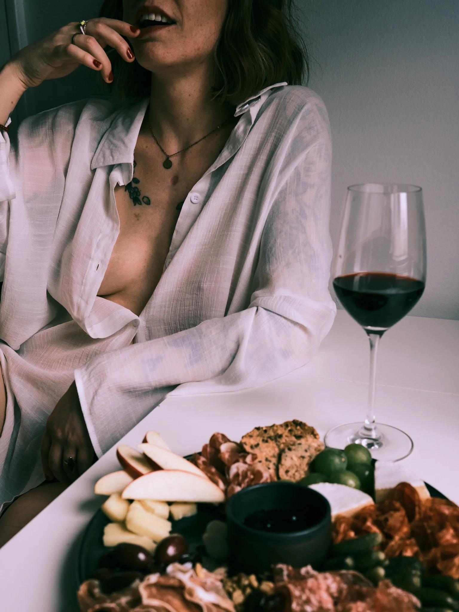 A woman with dark hair, wearing a light-colored, partially unbuttoned shirt, sitting at a table with a variety of foods and a glass of red wine.