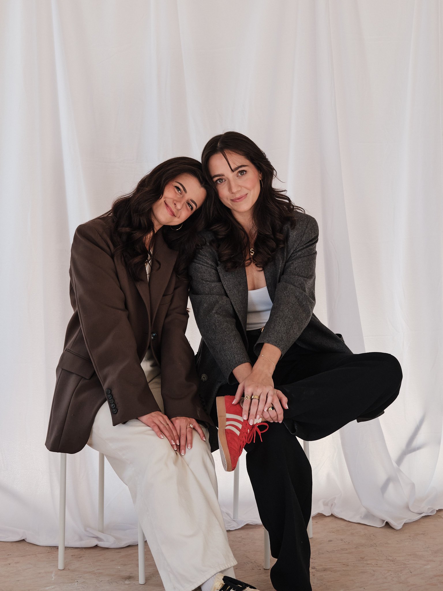 Two women sitting closely together, smiling at the camera, against a white backdrop. One is wearing a brown blazer and beige pants, the other a gray blazer and black pants, with red sneakers visible.