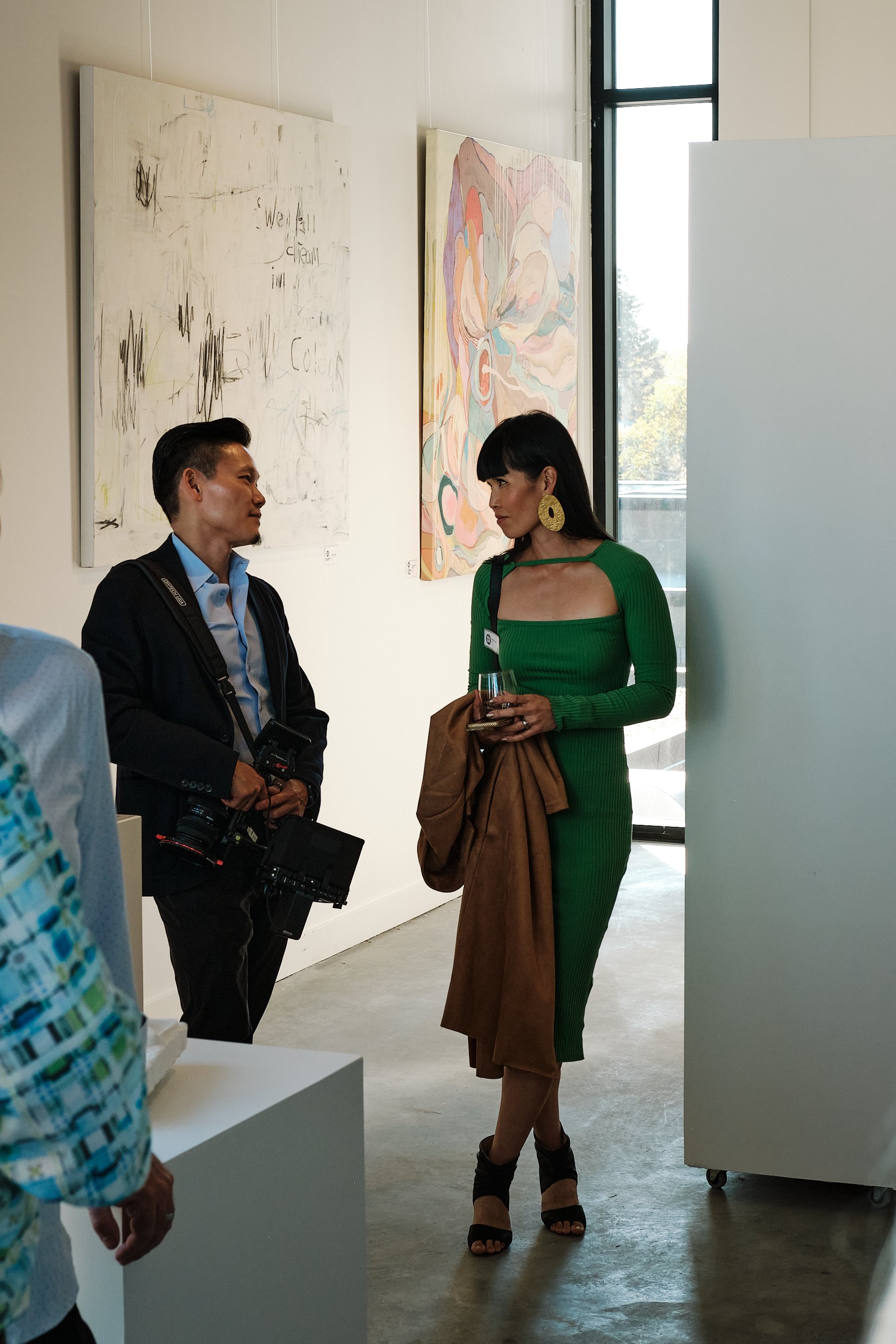 A woman in a green dress holding a glass, talking to a man with a camera at an art gallery. Taken in Edmonton, Alberta.