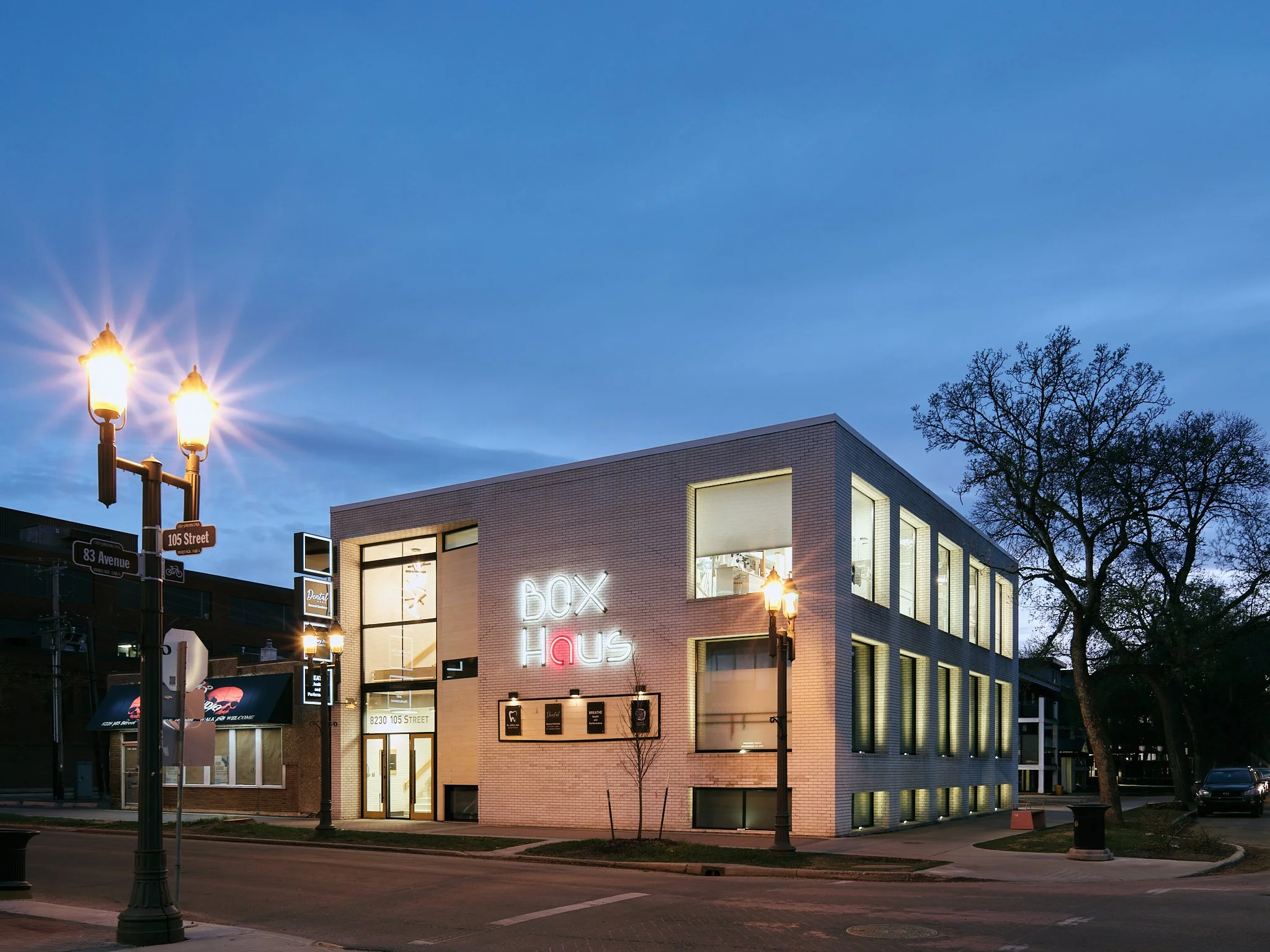 A modern, multi-story building with large windows and a sign that reads 'Box Haus,' illuminated at dusk. Street lamps and trees are visible in the foreground, and a clear evening sky is above. Box Haus off Whyte Avenue, Edmonton, Alberta.