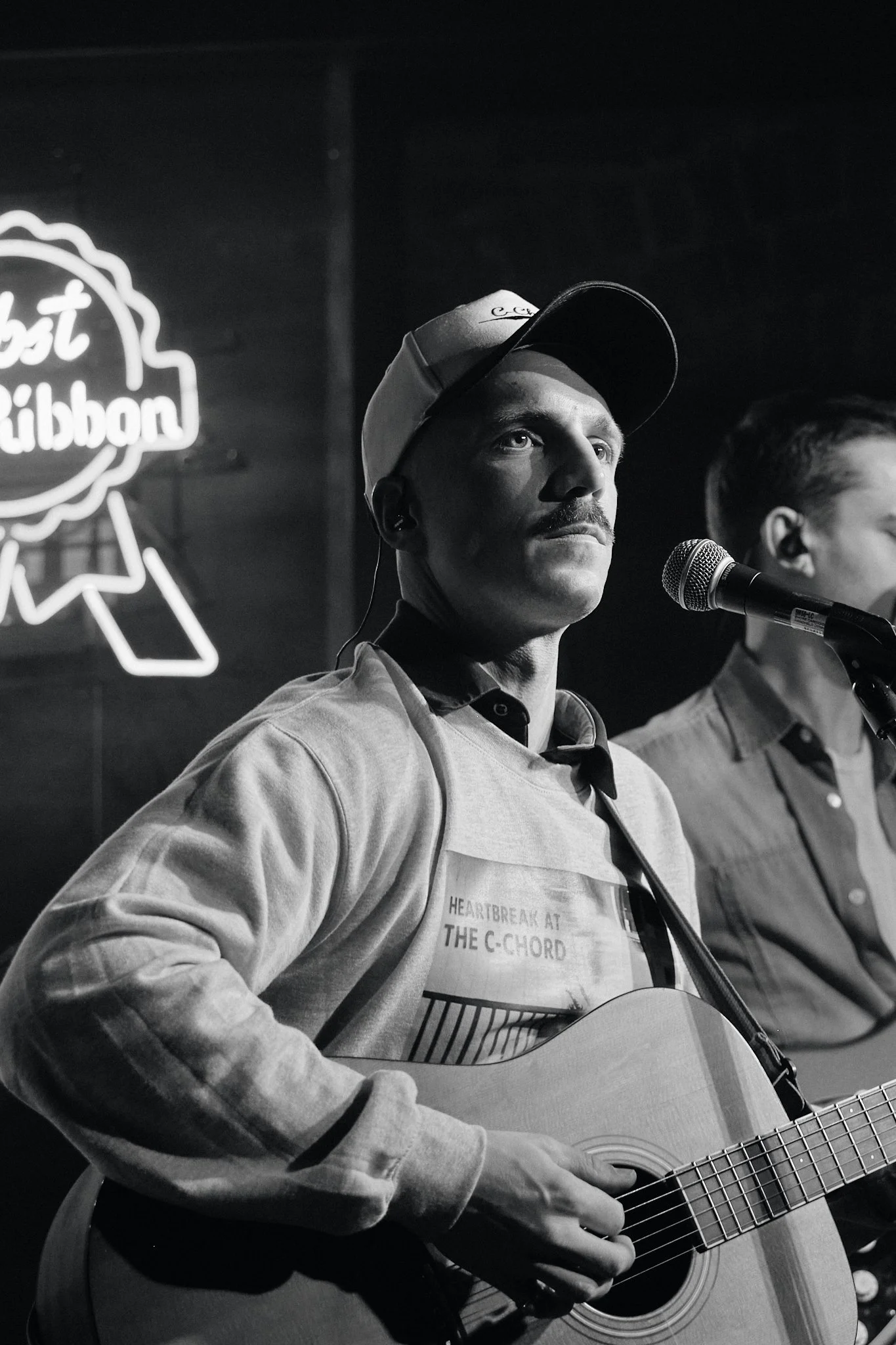 David Henneberg. Black and white photo of a musician wearing a white hat and a grey sweatshirt that says "Heartbreak in the C Chord" playing guitar and looking into the crowd, while playing at Dive Bar in Edmonton, Alberta.