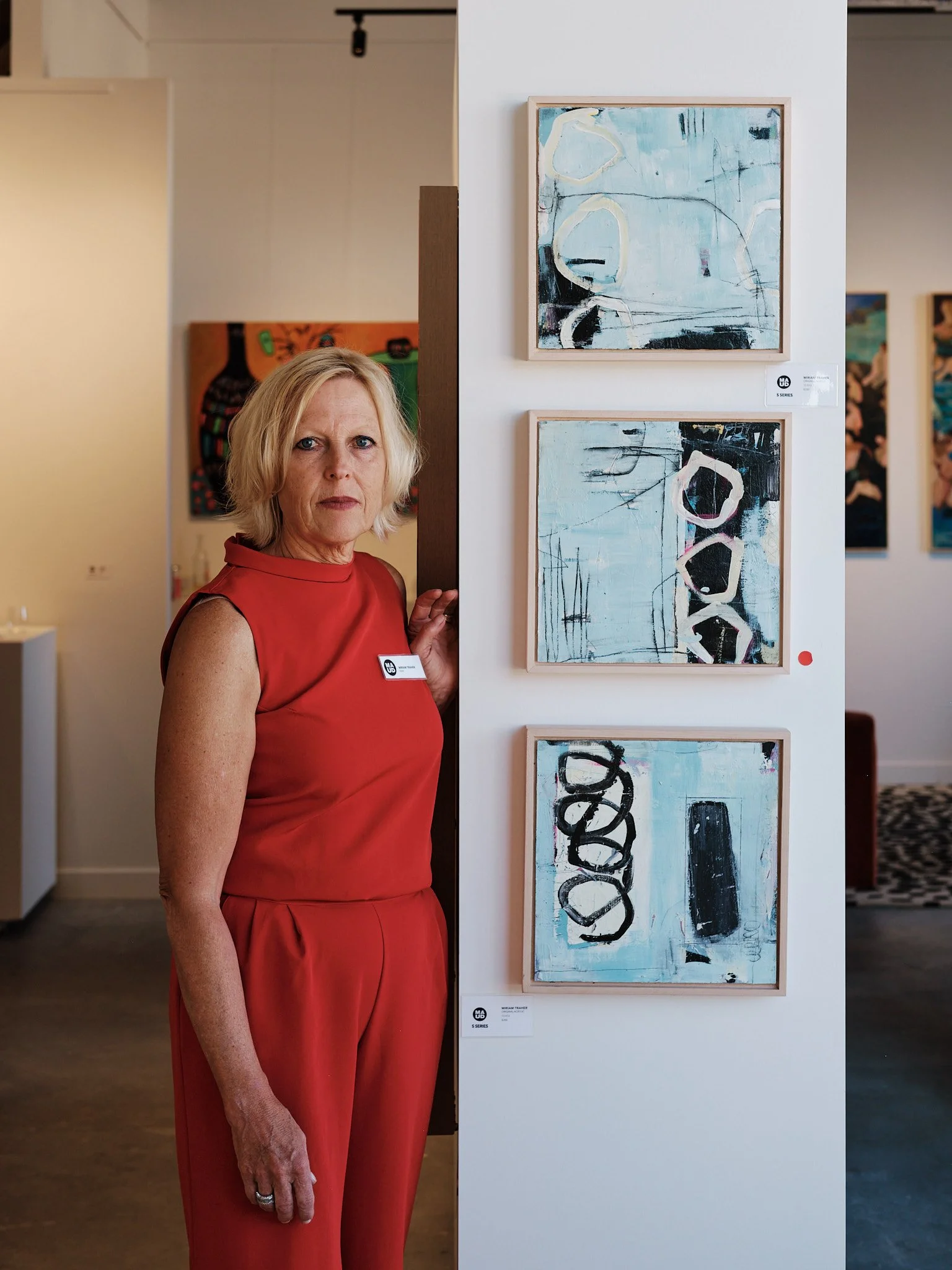 A woman with short blonde hair wearing a sleeveless red dress standing next to a white art display with four abstract paintings in a gallery. Edmonton, Alberta