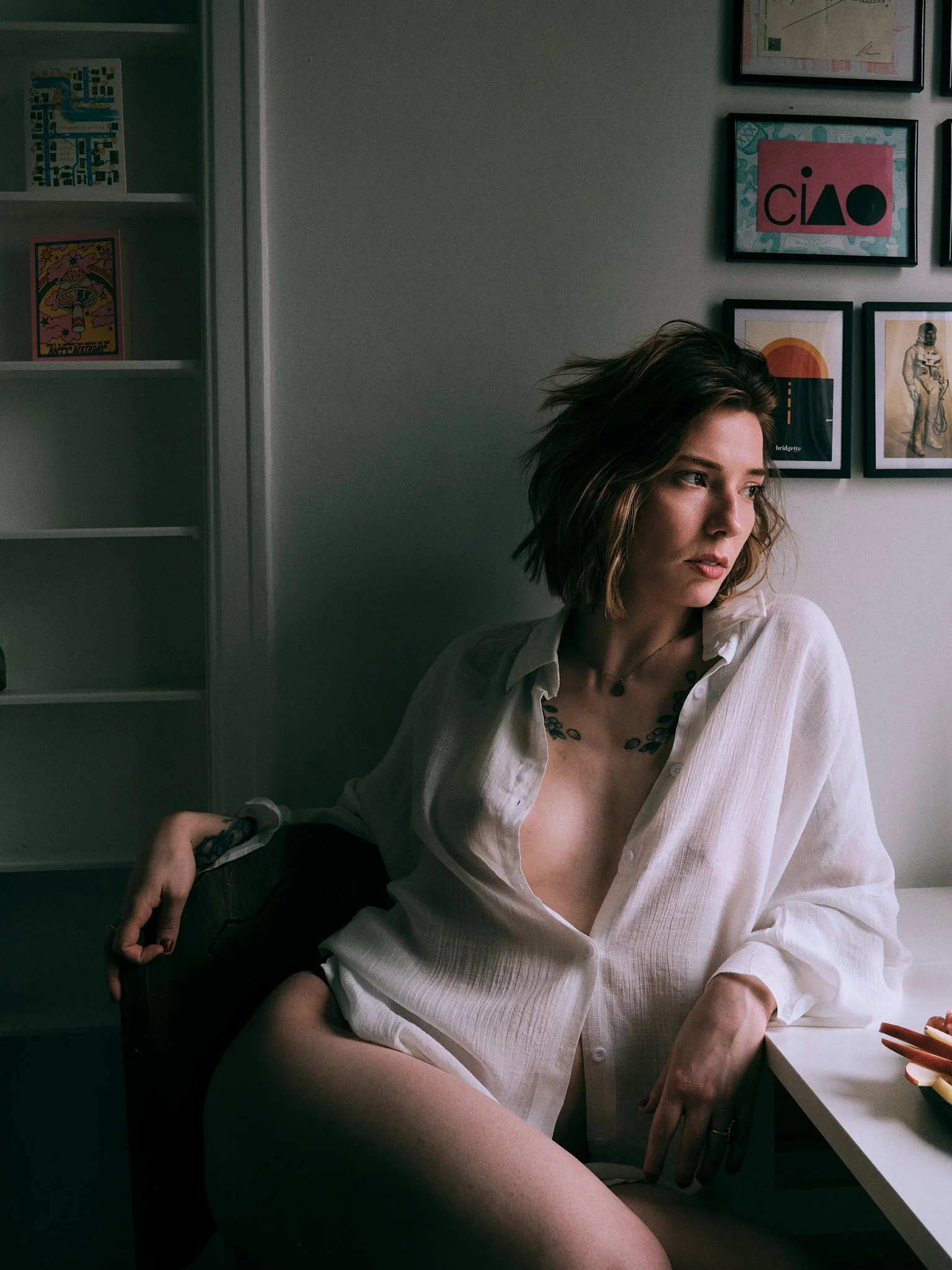 A woman with short wavy brown hair and light skin sitting at a desk with a white shirt unbuttoned, looking to her right. She is partially visible from the shoulders up. Behind her are framed pictures on the wall and a white bookshelf with colorful ca
