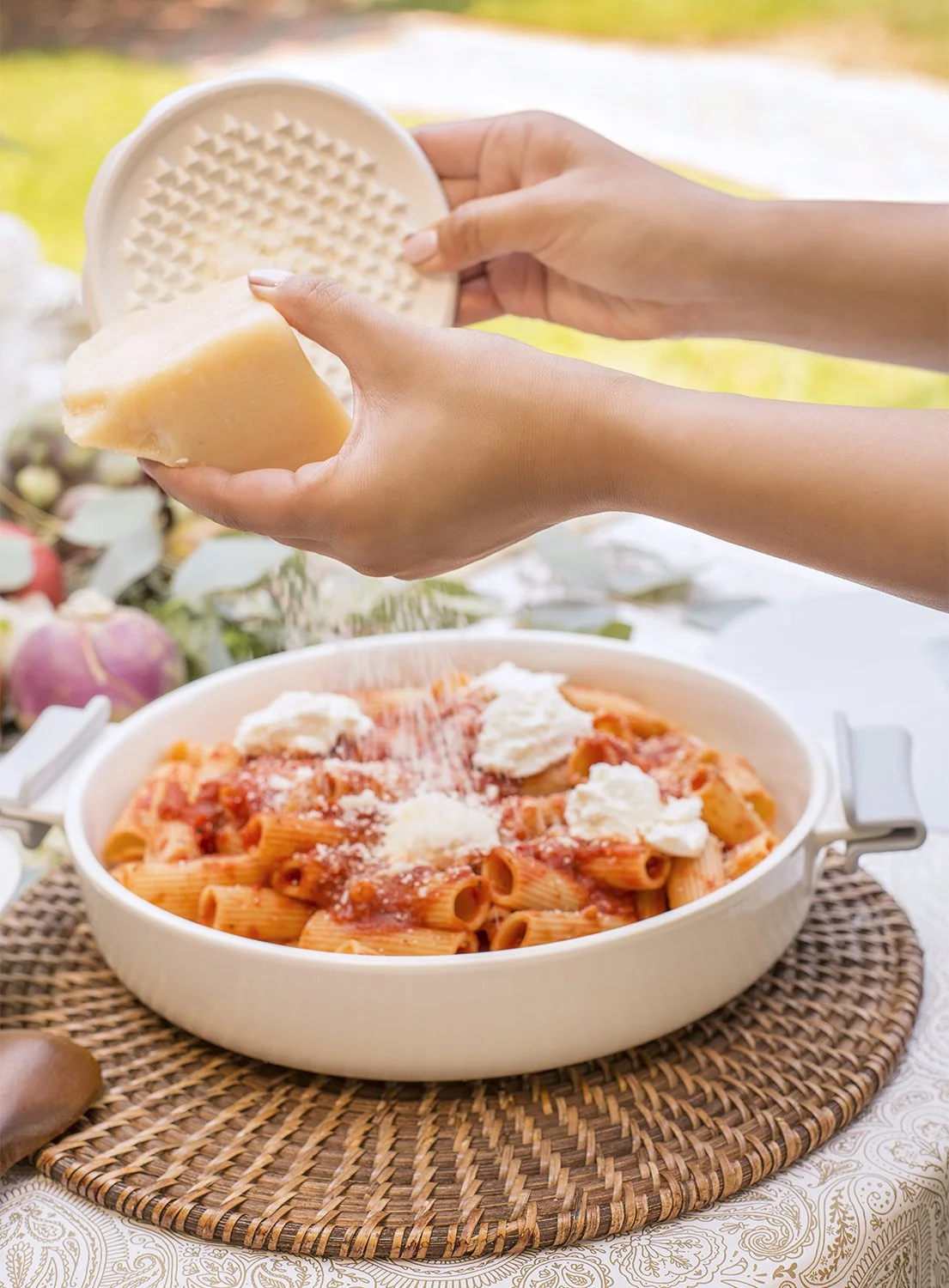 Hands grating cheese over a bowl of rigatoni pasta with tomato sauce and dollops of ricotta, set on a woven mat outdoors with a blurred background of greenery.