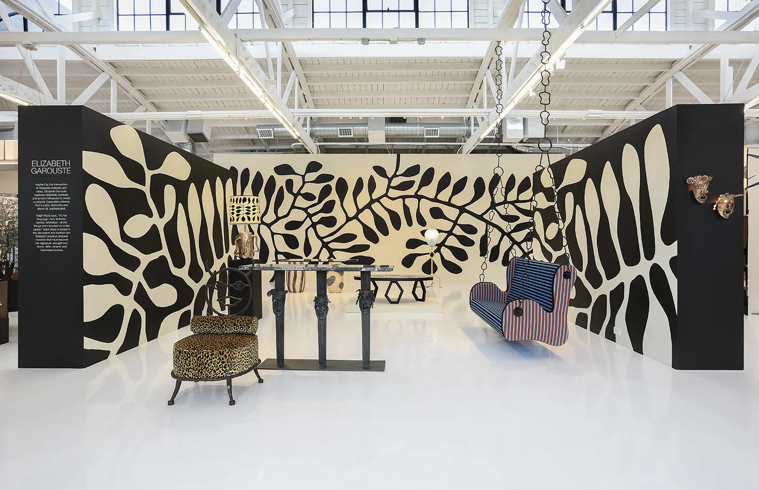 A modern art gallery, Ralph Pucci, features black and beige abstract leaf murals on the walls. The space includes various furniture pieces, such as a striped swinging couch, patterned chairs, and unique table designs, set on a polished white floor ag