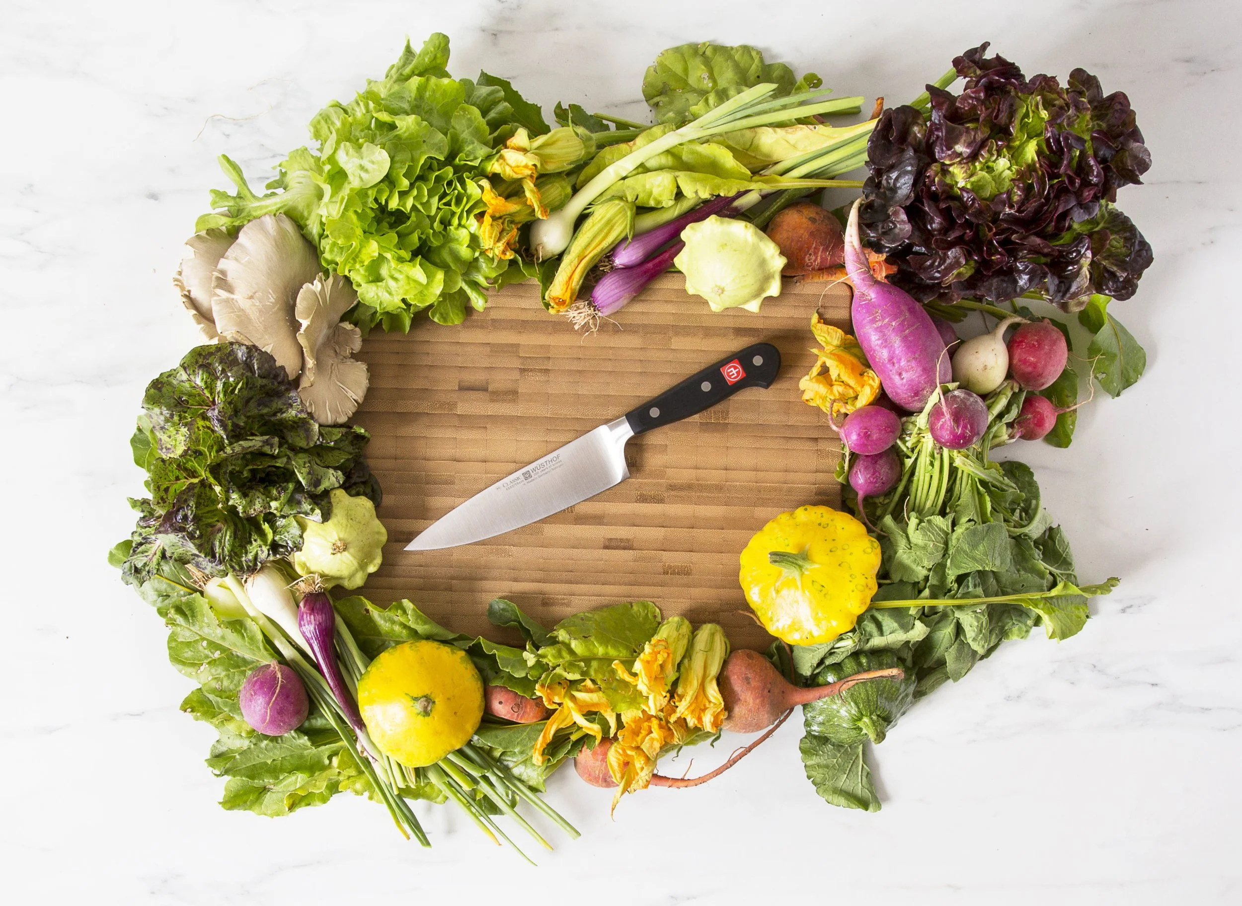 A variety of fresh vegetables, including lettuce, radishes, turnips, squash, and mushrooms, are arranged around a wooden cutting board. A chef's wusthof knife rests on the board, all set on a marble surface.