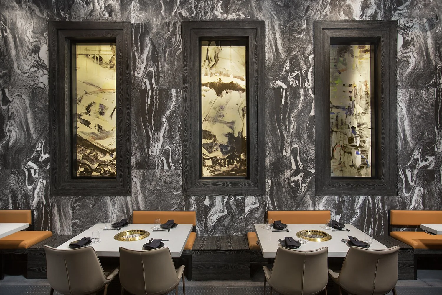 Interior of AB Steakhouse in Beverly Hills, with three tables, each equipped with built-in grills. The wall features a striking black and white marble pattern with three framed, abstract art pieces. The tables are set with dark napkins and surrounded