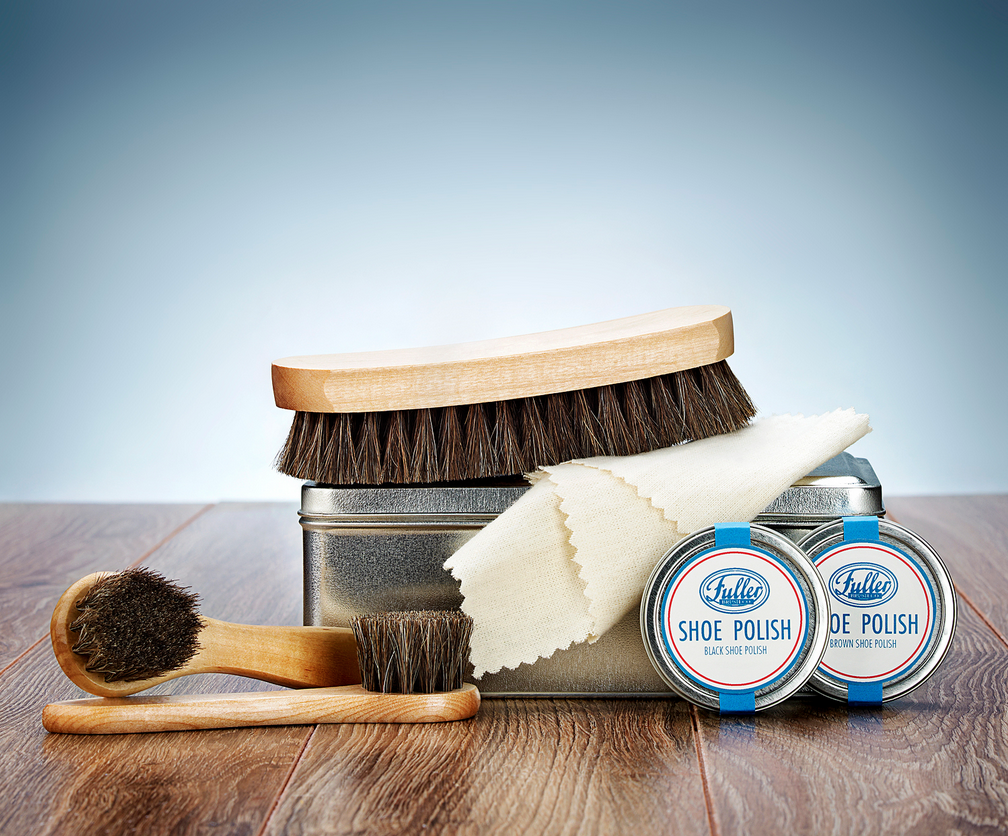A shoe care kit on a wooden surface, featuring two tins of shoe polish, a metal box, two shoe brushes with wooden handles, and a polishing cloth draped over the box. The background is a light, gradient blue.