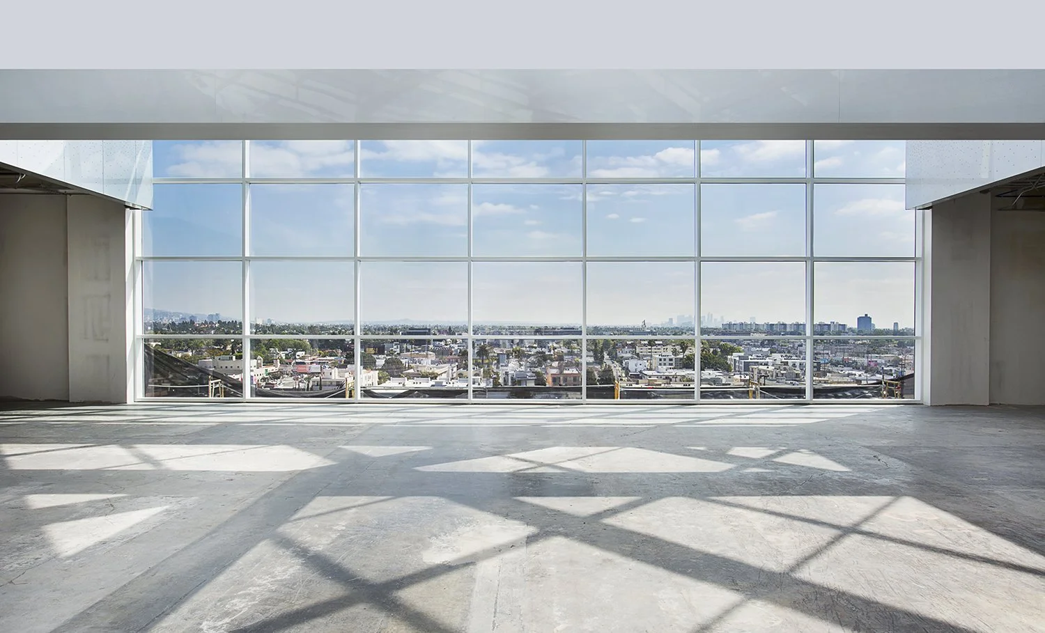 An empty interior space at the Beverly Center with a large grid of floor-to-ceiling windows, providing a panoramic view of Los Angeles with buildings and blue sky visible. The room has a concrete floor and is filled with natural light casting pattern