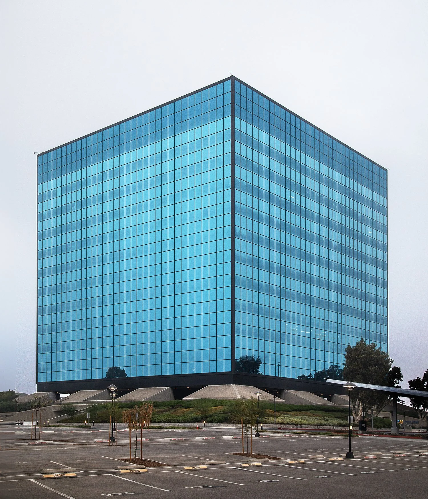 A large, modern cubic office building in Alhambra, CA, with reflective blue-tinted windows sits on a raised platform. The surrounding area is a mostly empty parking lot with sparse trees. The building is now owned by the Los Angeles of Department and