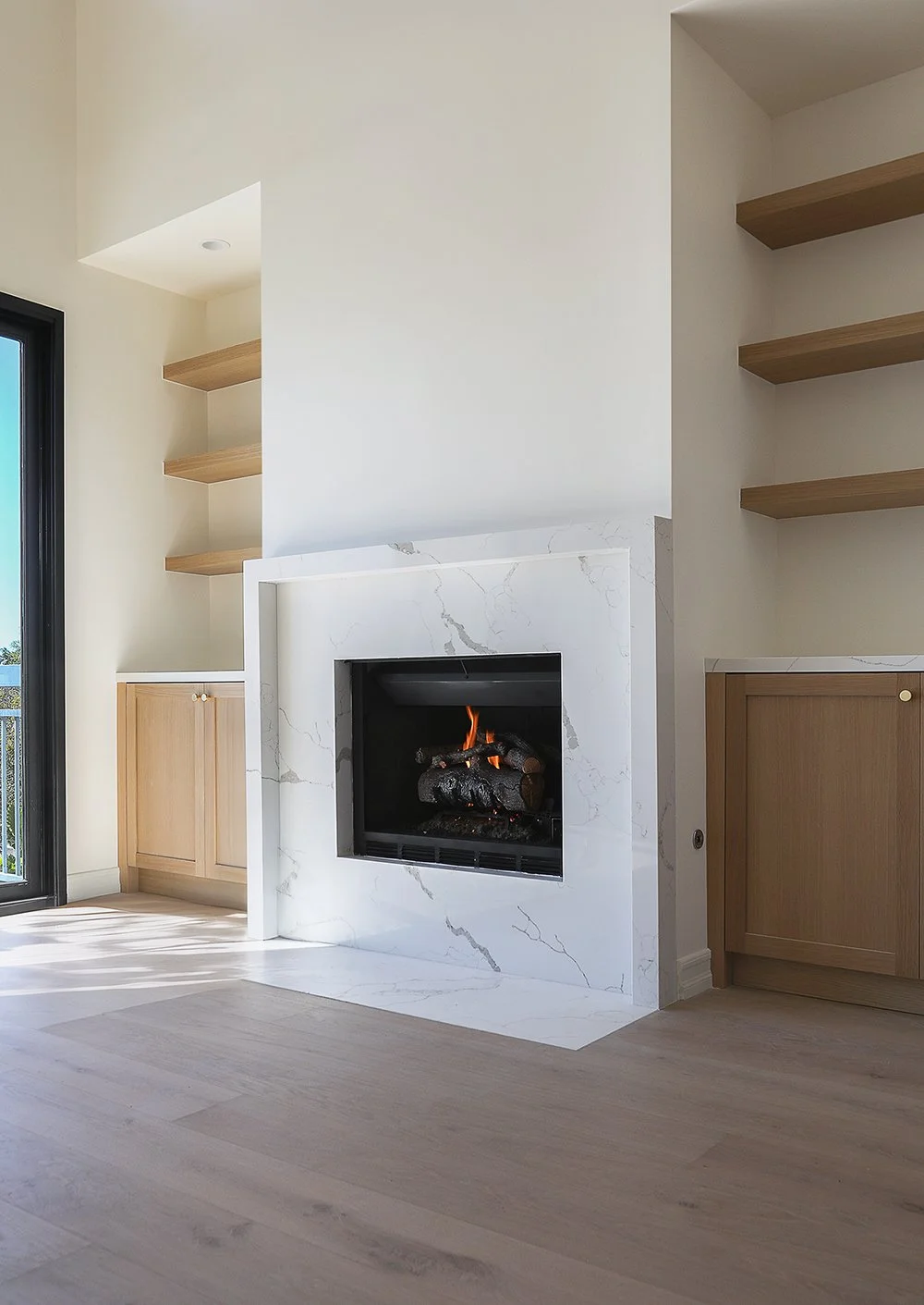 A modern living room features a sleek fireplace with a marble surround. The fire is lit, casting a warm glow. To the left and right of the fireplace are built-in wooden cabinets and open shelves. Sunlight streams in from a large window, illuminating 