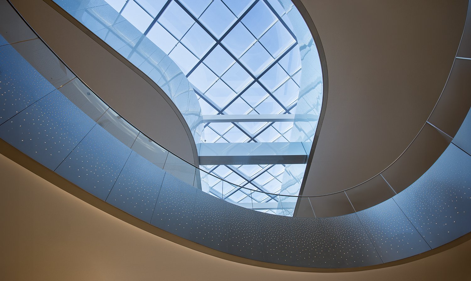 A view looking up at a modern architectural structure with a curved, glass ceiling and smooth, metallic railings. The ceiling is composed of large glass panels arranged in a grid pattern, allowing natural light to illuminate the interior space.