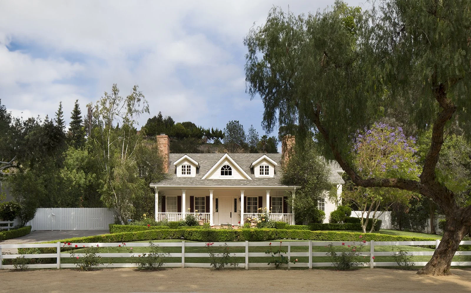 A charming, two-story house with a white picket fence and two large chimneys. The house features a front porch with columns, neatly trimmed bushes, and trees surrounding the property. There are three dormer windows on the roof, and the sky is partly 
