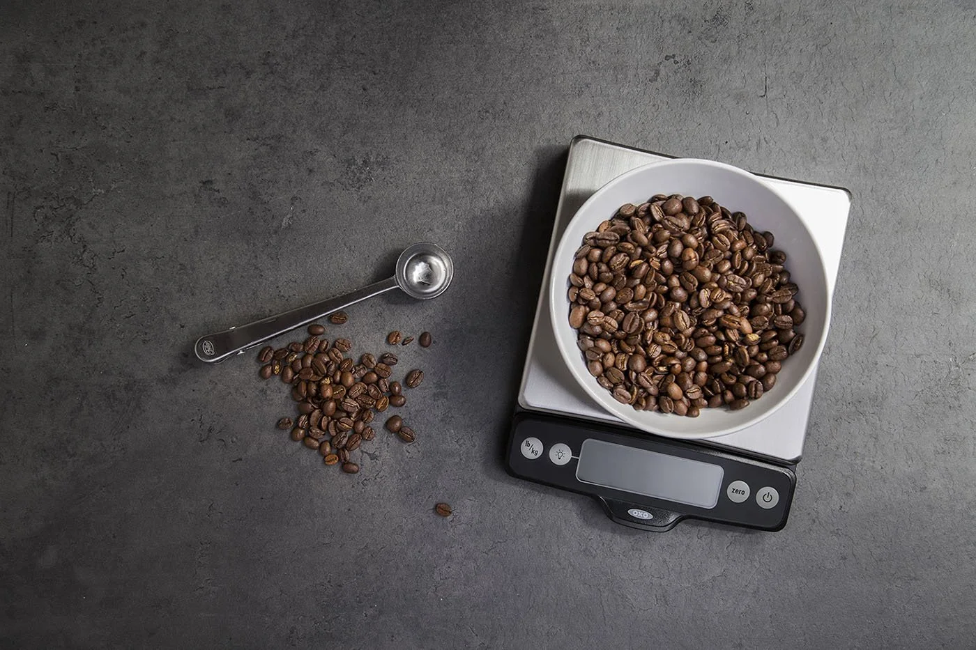 A bowl filled with coffee beans sits on a digital scale on a dark countertop. A metal scoop rests beside the scale, with a few coffee beans scattered around.