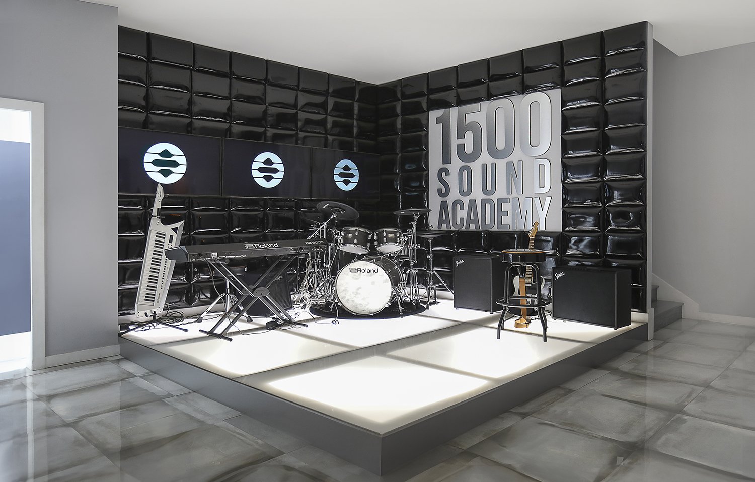 A modern music studio, called 1500 Sound Academy, owned by James Fauntleroy. The setup featuring a drum kit, keyboard, electric guitar, and microphone stand on a lit platform. The background has a wall with black acoustic panels, three screens with l