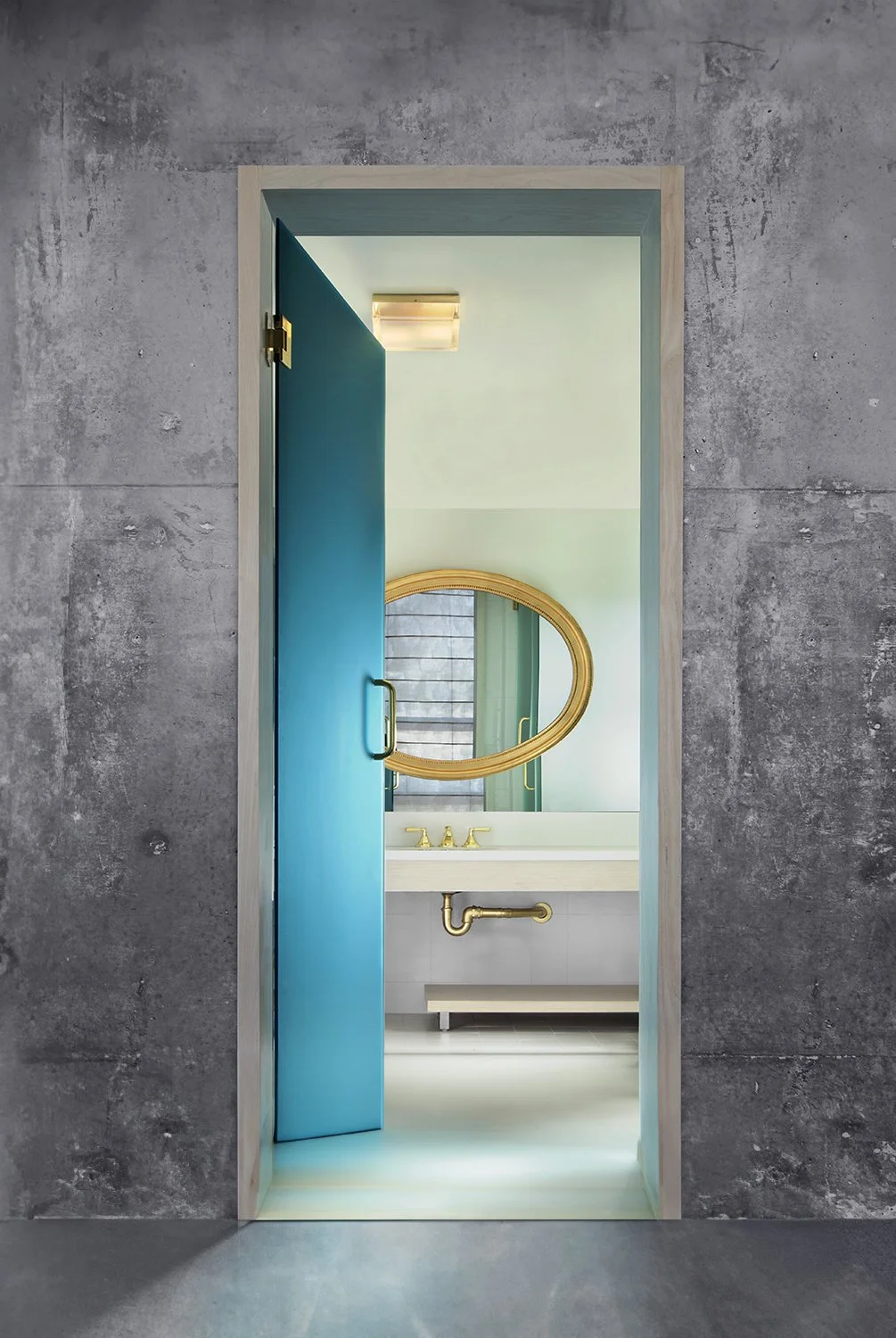 A modern bathroom designed by Knibb Design at the Line Hotel in Los Angeles, CA. A blue door slightly ajar, revealing a white sink, a brass faucet, and a large round gold-framed mirror. The room is accented with muted colors and illuminated by a ceil