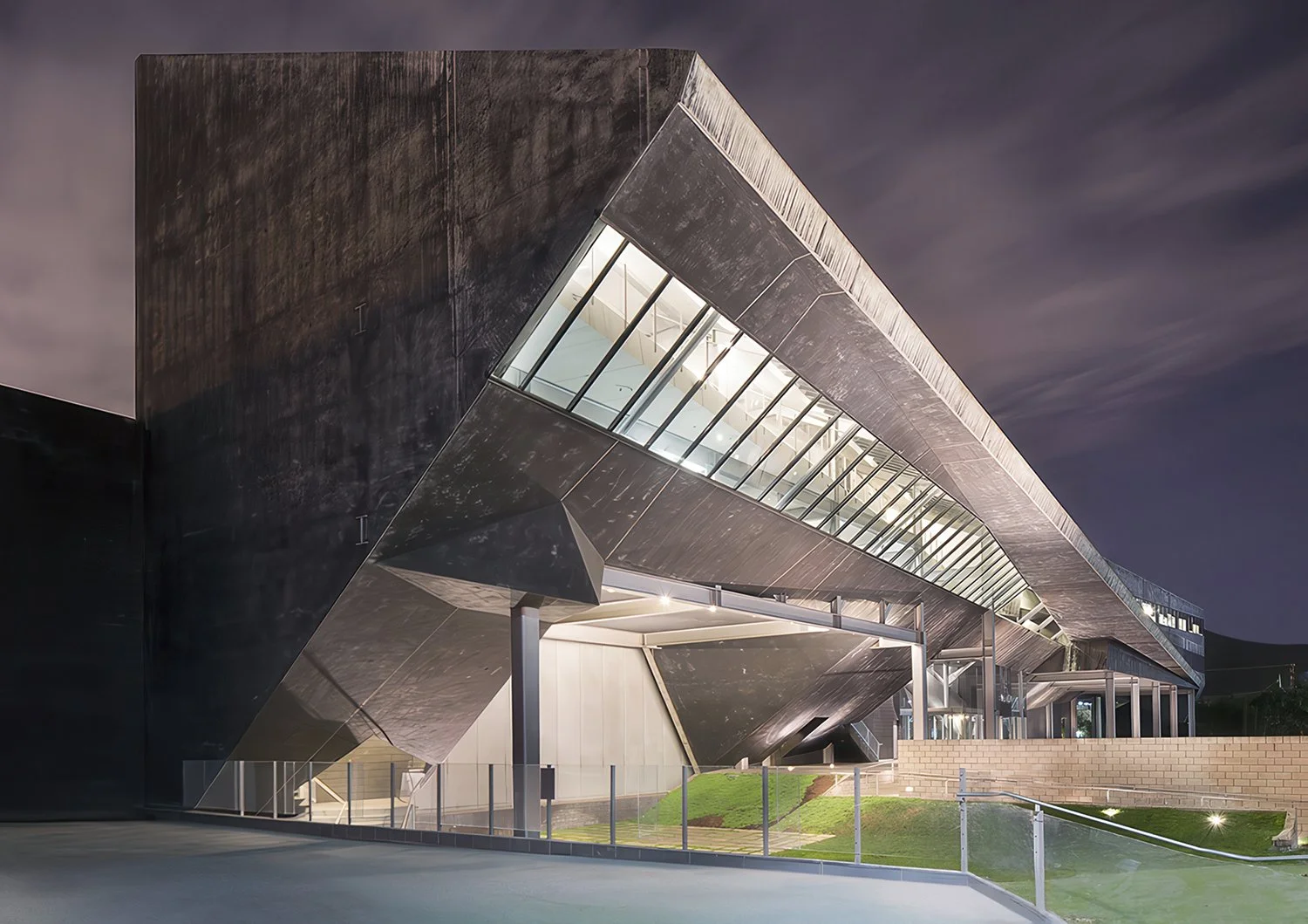 A modern, angular building with sharp geometric lines illuminated at night. The structure features dark, textured surfaces with large, slanted windows and an overhanging roof. It stands on a landscaped area with glass railings and outdoor lighting.