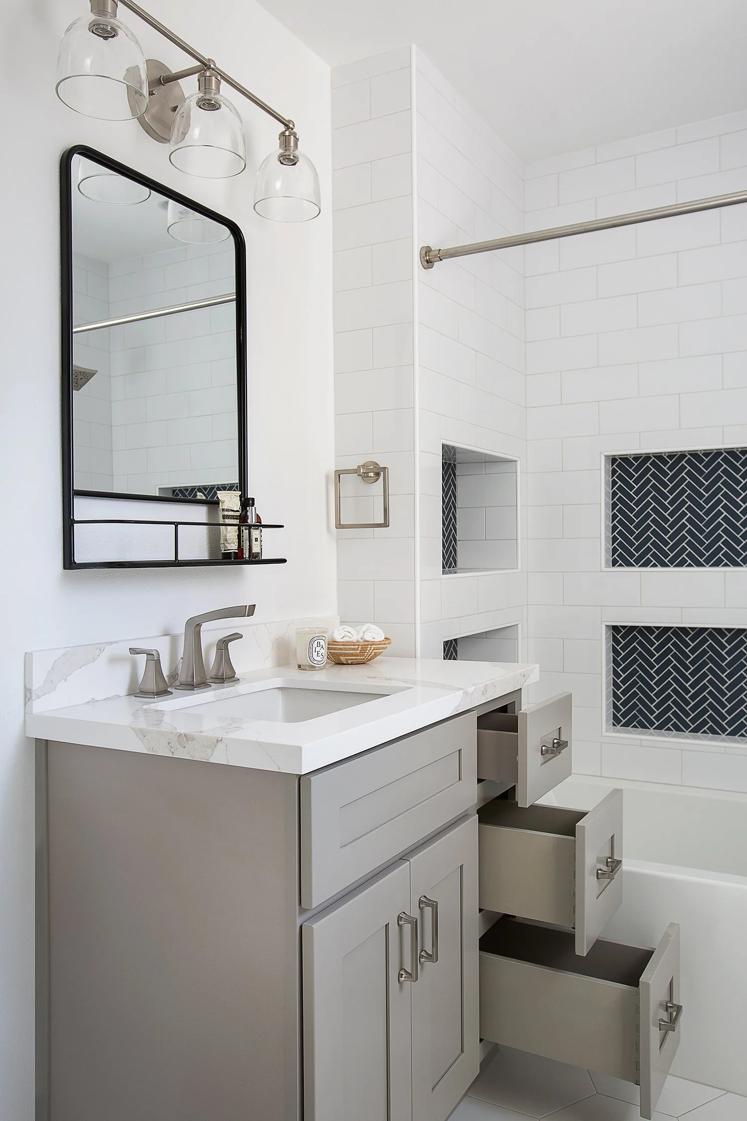 A modern bathroom with white subway tile walls and dark blue herringbone accents. The vanity has a marble countertop, a rectangular mirror with a black frame, and three open drawers. Above the mirror is a three-light fixture. The bathtub has a chrome