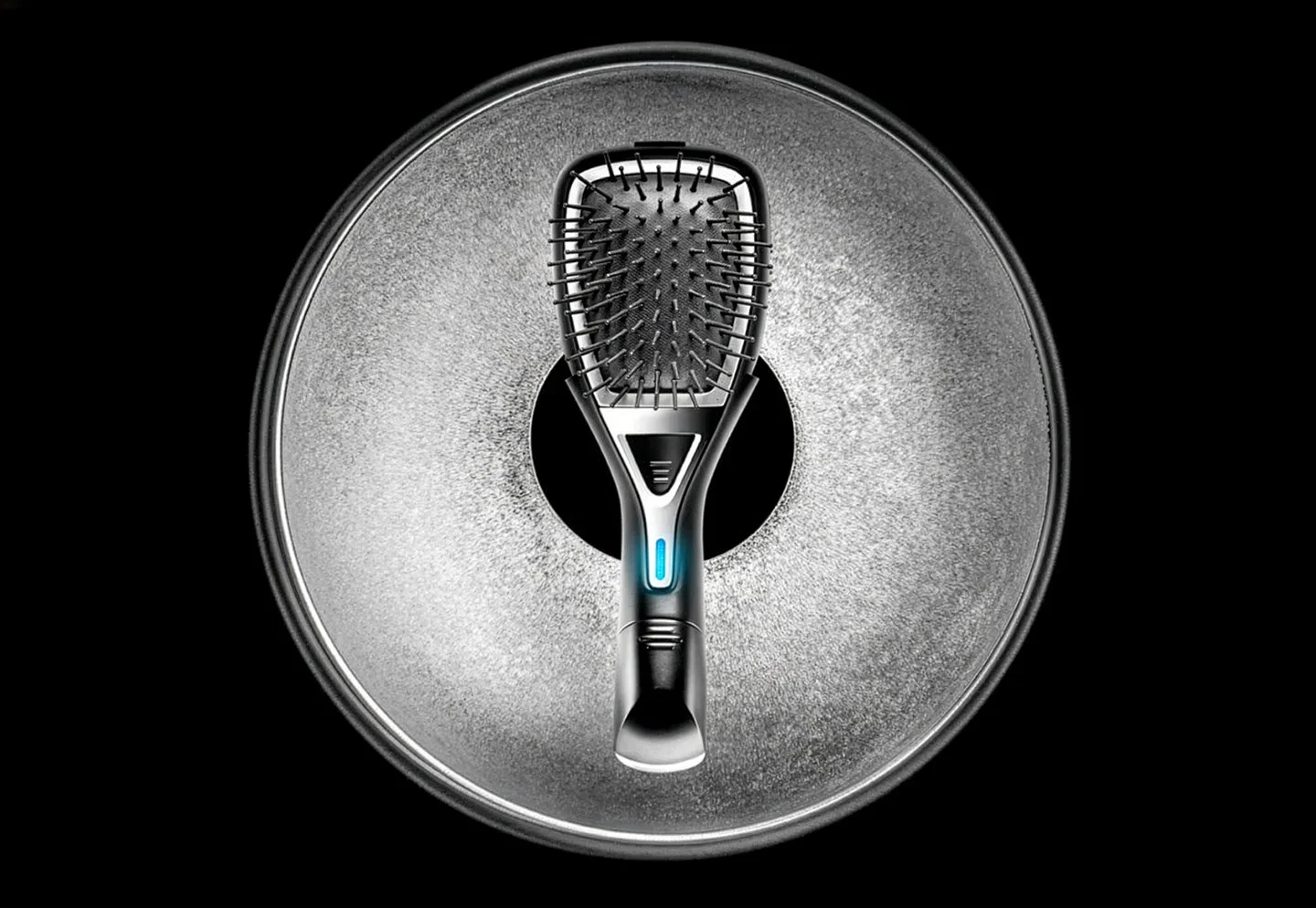 A modern metallic hairbrush with a blue accent is standing upright in the center of a round, textured silver background with a circular cut-out, all set against a black backdrop.