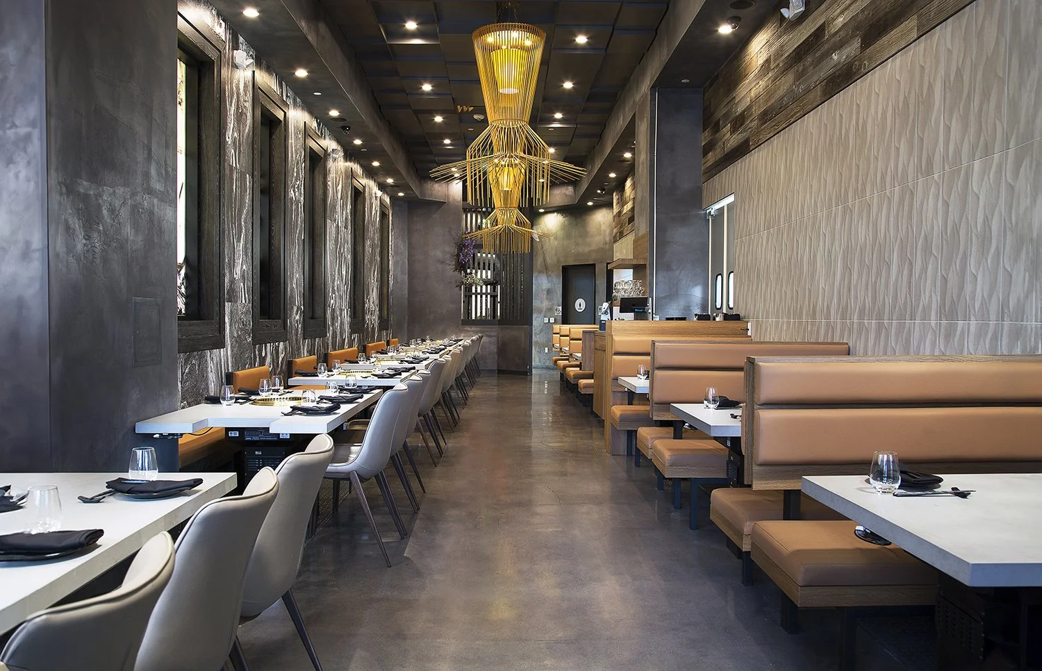 AB Steakhouse is a modern, sleek restaurant by Chef Akira Back. It has a polished concrete floor, lined with beige booth seating on the right and white table-chairs on the left. Two large, decorative chandeliers hang from the ceiling, and the space f