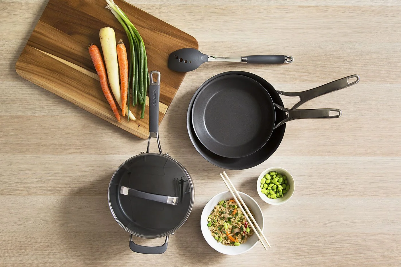 Cooking utensils on a wooden table: two black frying pans, a pot with a lid, and a spatula. A wooden board with carrots and green onions, and bowls of mixed vegetables and edamame are nearby. Chopsticks rest on one bowl.