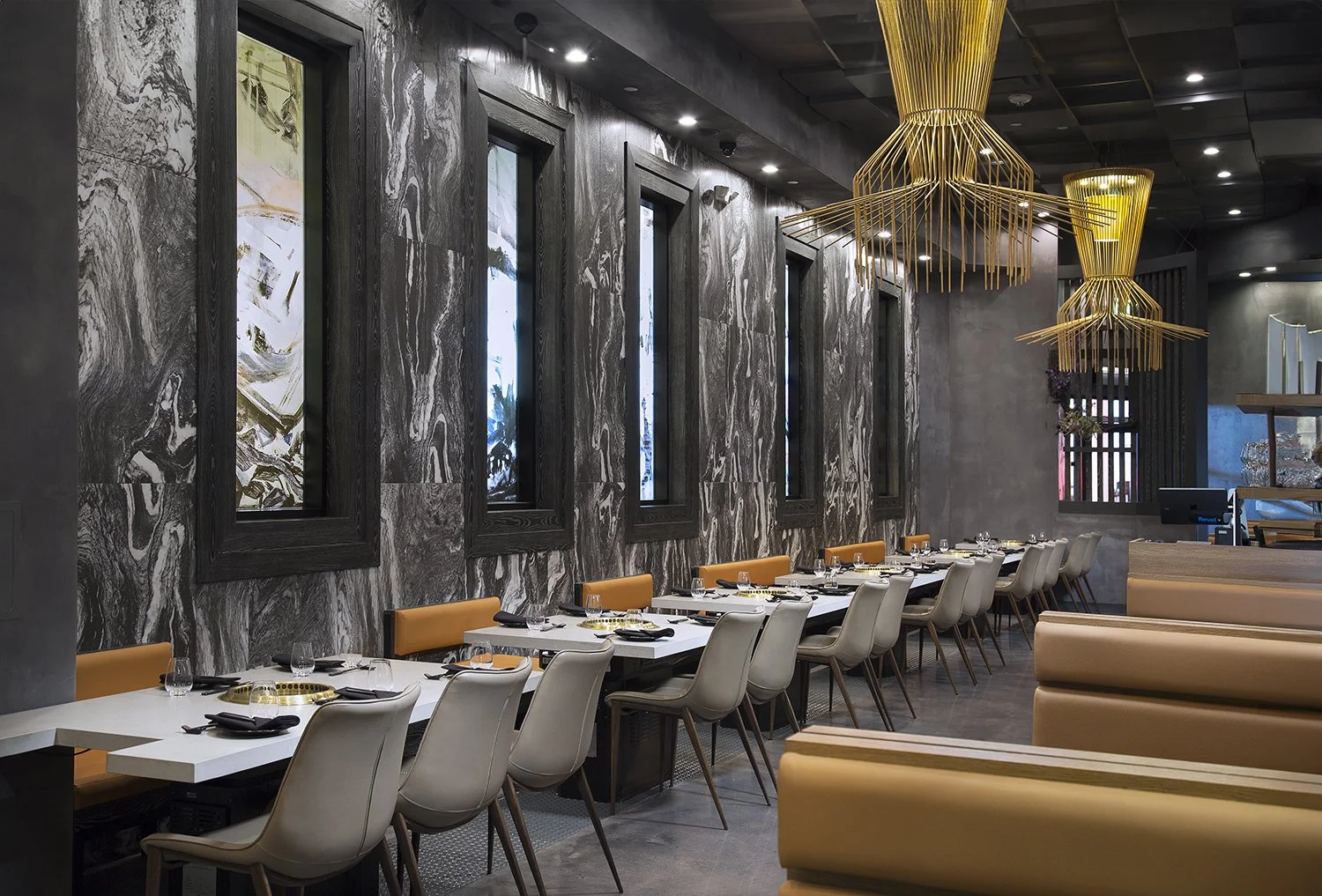 AB Steakhouse is a modern, elegantly designed restaurant interior featuring long tables with white tabletops and a mix of beige and gray chairs. The walls are adorned with marble patterns and framed artwork. The ceiling boasts artistic, golden light 