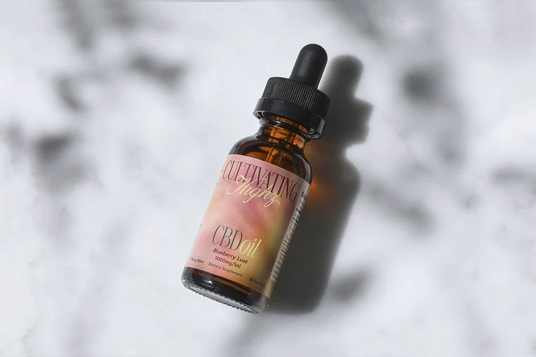 A bottle of CBD oil labeled "Cultivating Magic" with a dropper lid lies on a light marble surface. The label features a gradient design from maroon to peach, with shadow patterns in the background.