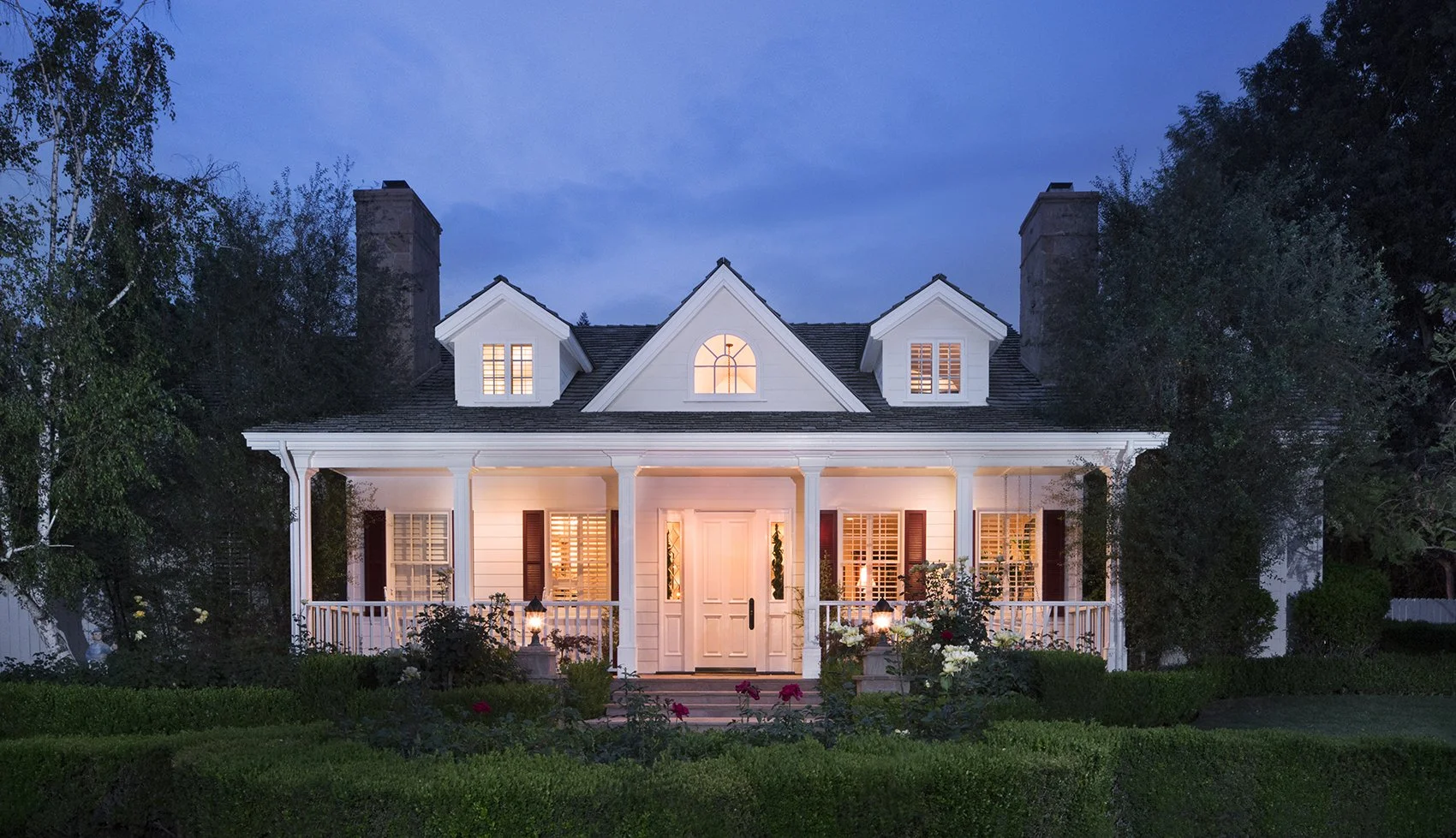 A charming, white, two-story house illuminated at dusk. The house features a porch with columns, dormer windows on the roof, and a well-manicured front yard with hedges and rose bushes. Two chimneys are visible on either side of the house.
