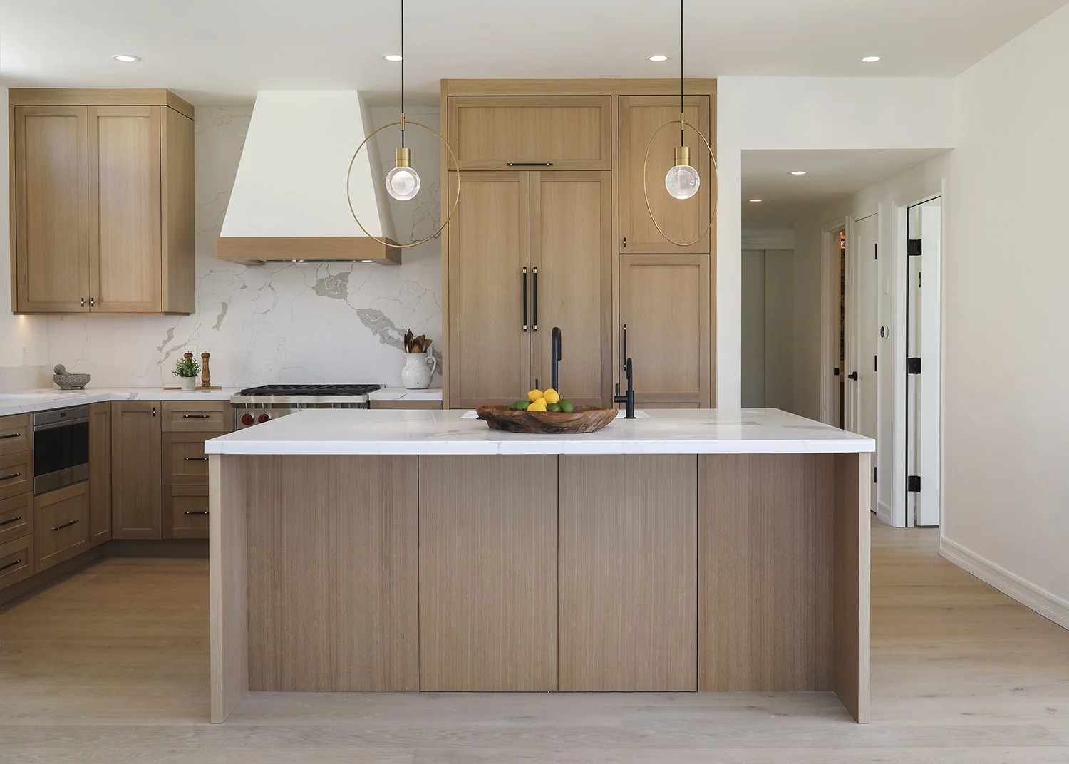Modern kitchen with light wood cabinets and a white marble backsplash. A large island with a white countertop holds a bowl of lemons. Pendant lights hang above the island, and built-in appliances are visible. The floor is light wood.