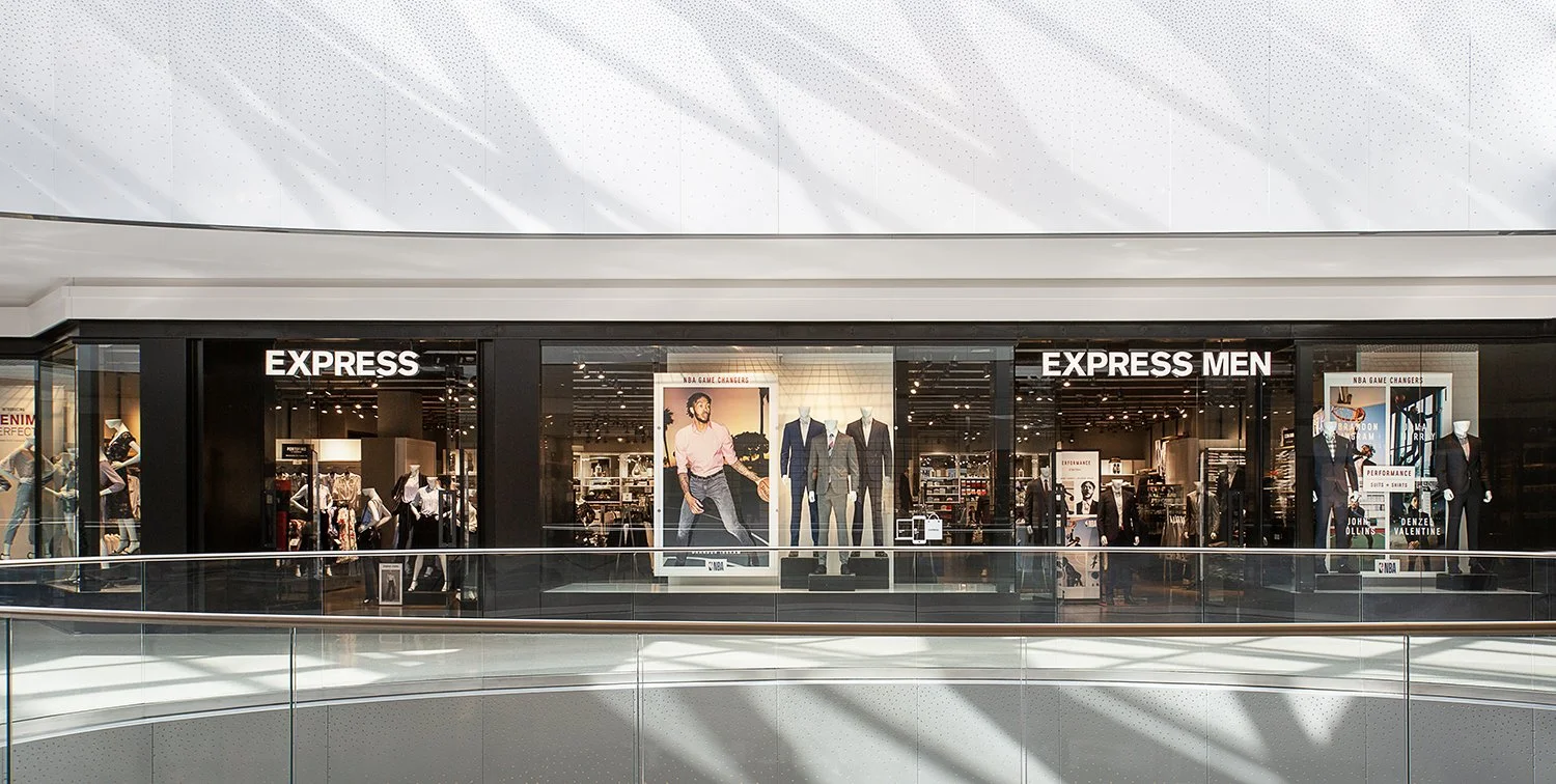 Front view of a clothing store. The store is named "EXPRESS" with sections for general apparel and a specific "EXPRESS MEN" area. The interior is modern with various clothes on display and large posters featuring men in stylish outfits. Shoppers are 