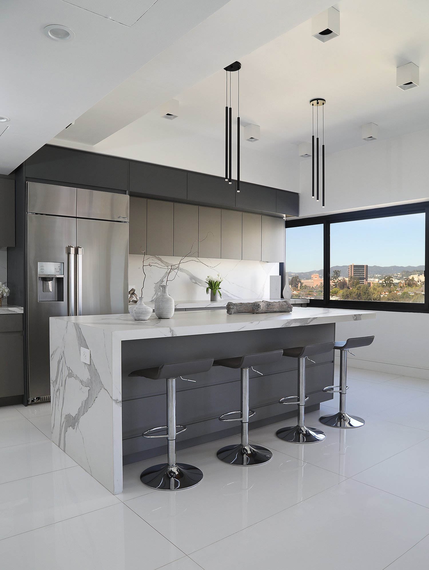 A modern kitchen with sleek design features a large island with a marble countertop and four black barstools. Stainless steel appliances, including a refrigerator, are integrated into the dark cabinetry. Large windows provide a scenic view outside.