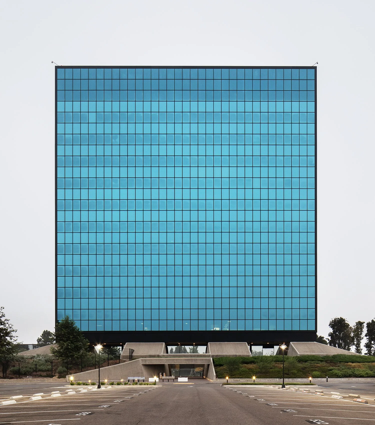 A large, modern cubic office building in Alhambra, CA, with reflective blue-tinted windows sits on a raised platform. The surrounding area is a mostly empty parking lot with sparse trees. The building is now owned by the Los Angeles of Department and