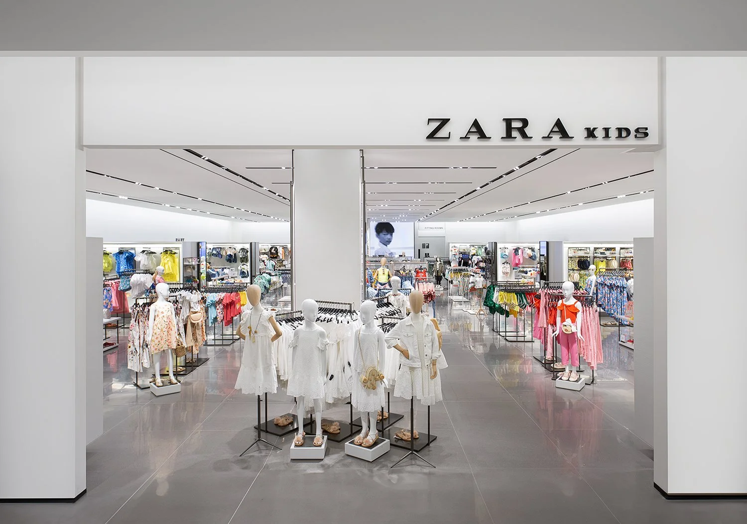 A brightly lit Zara Kids store with neatly arranged children's clothing displays. Mannequins wearing various outfits are at the entrance. Racks of colorful clothing are organized on the sides and in the background. The store has a modern and spacious