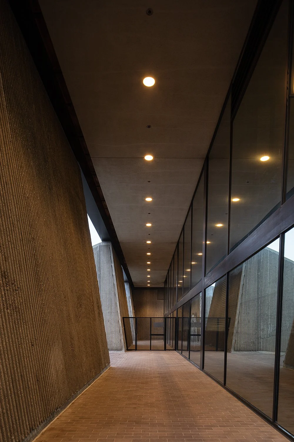 A modern architectural walkway featuring a textured, angled concrete wall on one side and a wall of large reflective glass panels on the other. The ceiling is lined with recessed lights, creating a warm and inviting ambiance.
