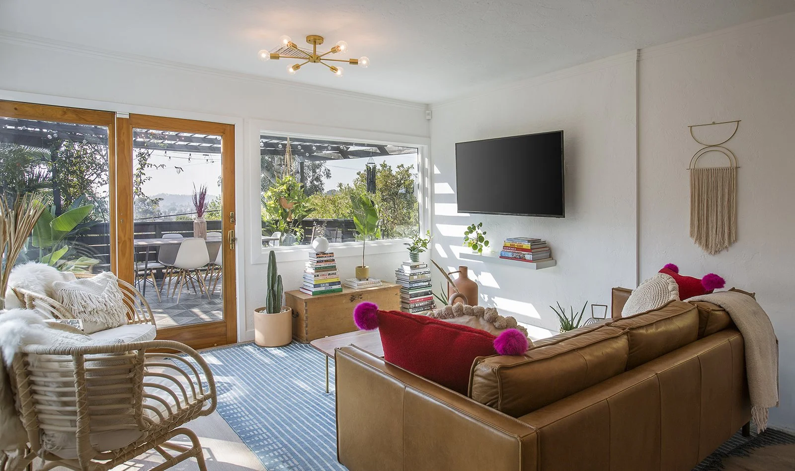 A bright and cozy living room featuring a brown leather sofa with pink pom-pom cushions, a rattan chair, a wall-mounted TV, and a stack of books. Glass doors open to a balcony with outdoor seating and greenery. Minimalist decor includes hanging plant