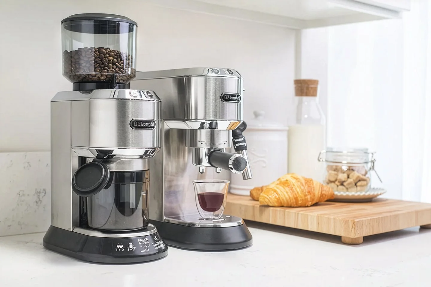A stainless steel coffee grinder and espresso machine sit on a kitchen counter. The grinder is filled with coffee beans, and the machine dispenses espresso into a small glass. Nearby are a croissant, milk, and a jar of sugar cubes.