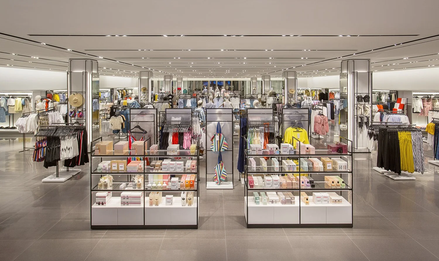 A spacious and well-lit clothing store, Zara,  with multiple sections. The central area features shelves displaying skincare and cosmetic products. The surrounding racks display various clothing items. The store has a sleek, modern design with a gray