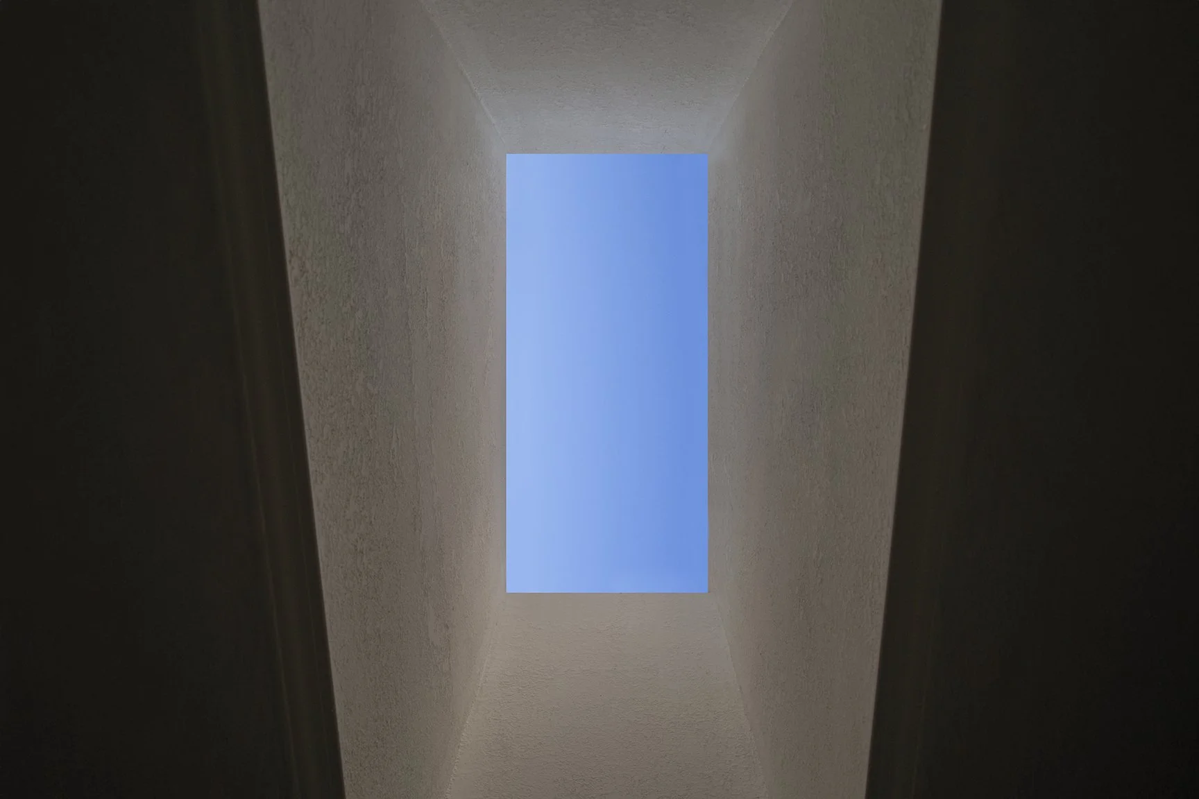 A rectangular skylight framed by textured white walls opens up to a clear blue sky, viewed from below within a narrow, vertical shaft-like space.