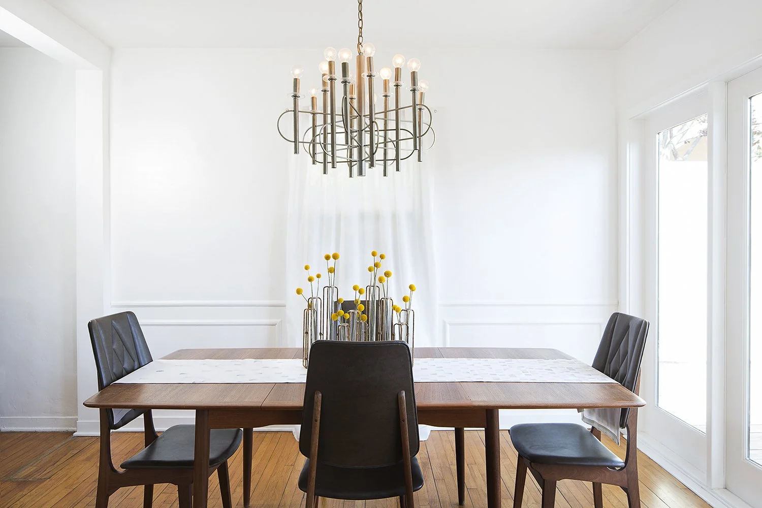 A modern dining room with a wooden table, four black chairs, a white runner, and a centerpiece with yellow flowers in metal vases. Above the table hangs a large, circular, multi-bulb chandelier. The room is well-lit with white walls and a floor-to-ce