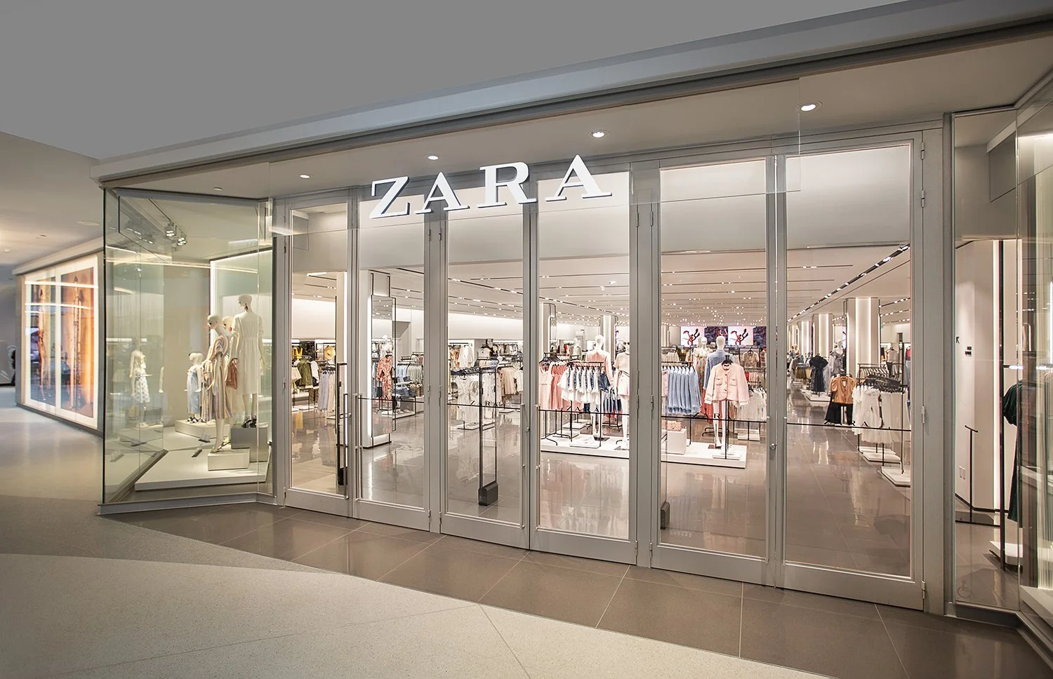 A storefront view of a Zara retail clothing store in a shopping mall. The entrance features glass doors, mannequins dressed in fashionable outfits on the left, and a well-lit interior displaying various clothing items on racks.