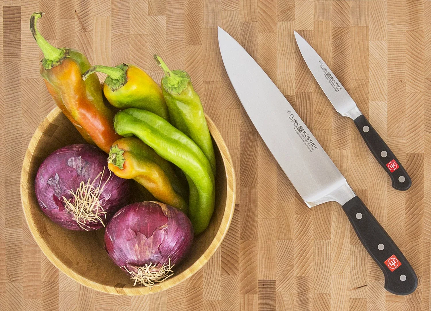 A wooden bowl with red onions and green peppers is on a wooden countertop. Beside the bowl, there are two wusthof knives with black handles placed parallel to each other.