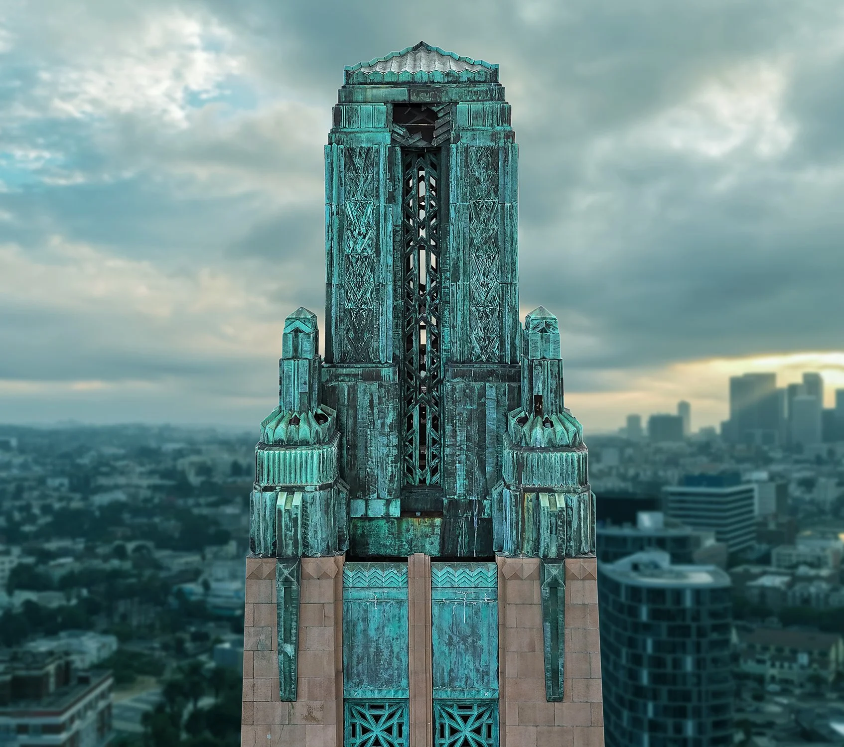 A close-up of an ornate, greenish-bronze art deco tower against a cloudy sky. It is called the Bullocks Building and is located in Los Angeles. The detailed architectural design stands out, with a city skyline and tall buildings visible in the backgr