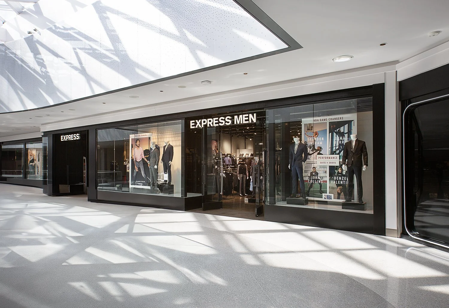 Storefront of Express Men at a shopping mall, featuring mannequins dressed in formal and casual attire displayed in large glass windows. The store's name is prominently visible above the entrance. The brightly lit mall environment creates reflections