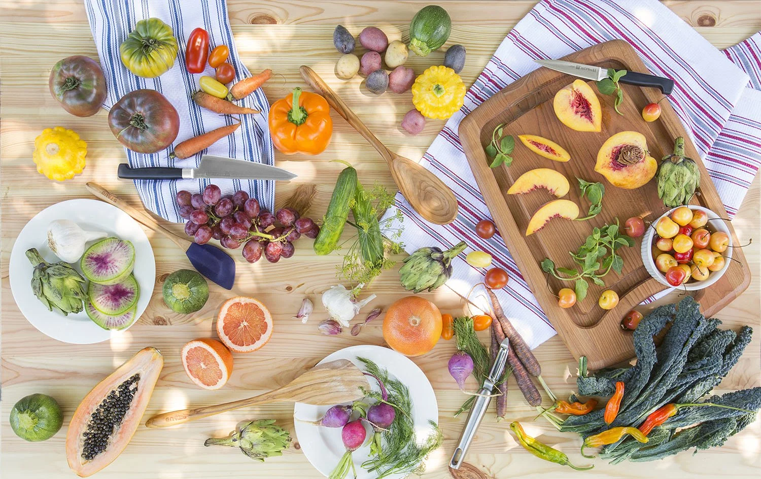 A colorful assortment of fresh fruits and vegetables on a wooden table. Includes peaches, grapes, peppers, artichokes, citrus, papaya, and leafy greens. A cutting board with sliced produce and knives is also visible.
